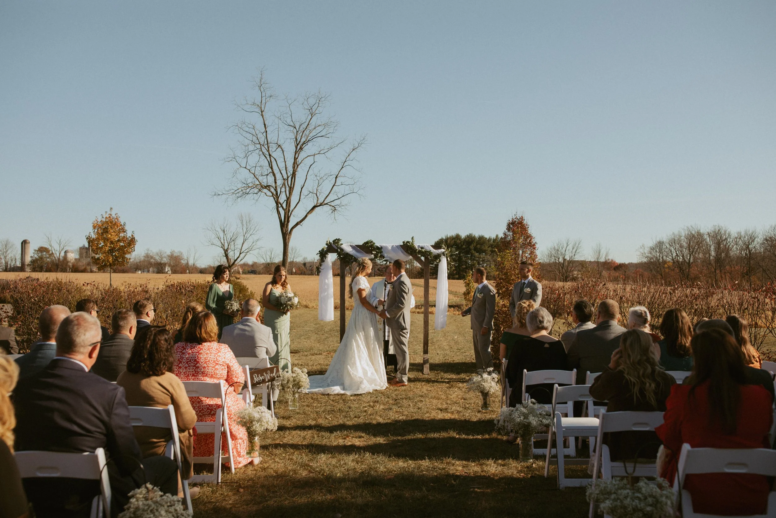 A wedding ceremony outdoors with a couple exchanging vows under a small decorated arch, surrounded by guests seated in white chairs on a grassy field with trees and a clear sky in the background.