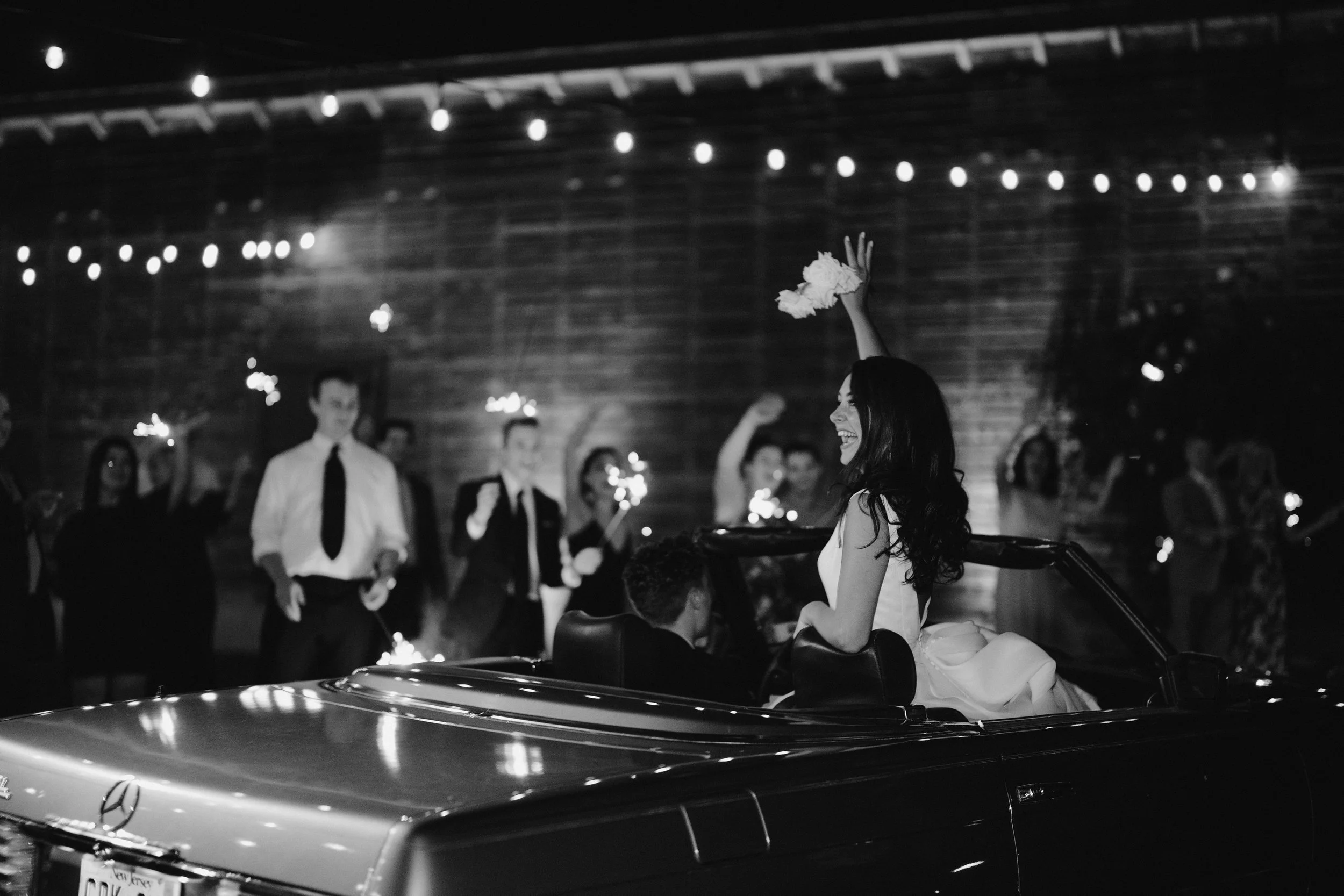 A joyful woman in a wedding dress holding a bouquet, celebrating at a wedding reception with friends and family in a rustic wooden venue illuminated with string lights, with a vintage car in the foreground.