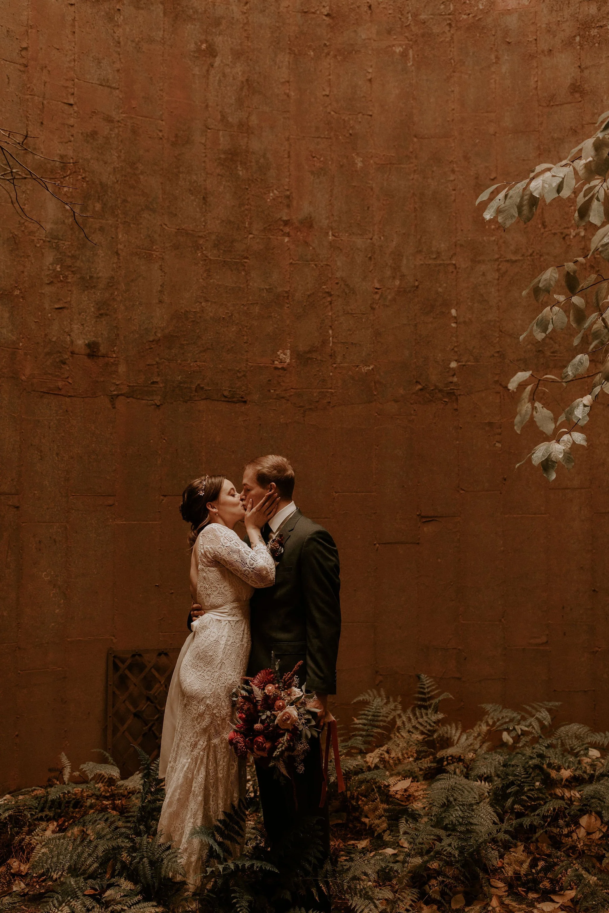 A bride and groom sharing a kiss during their wedding ceremony, standing closely together with a large rustic brick wall behind them, surrounded by plants and foliage.