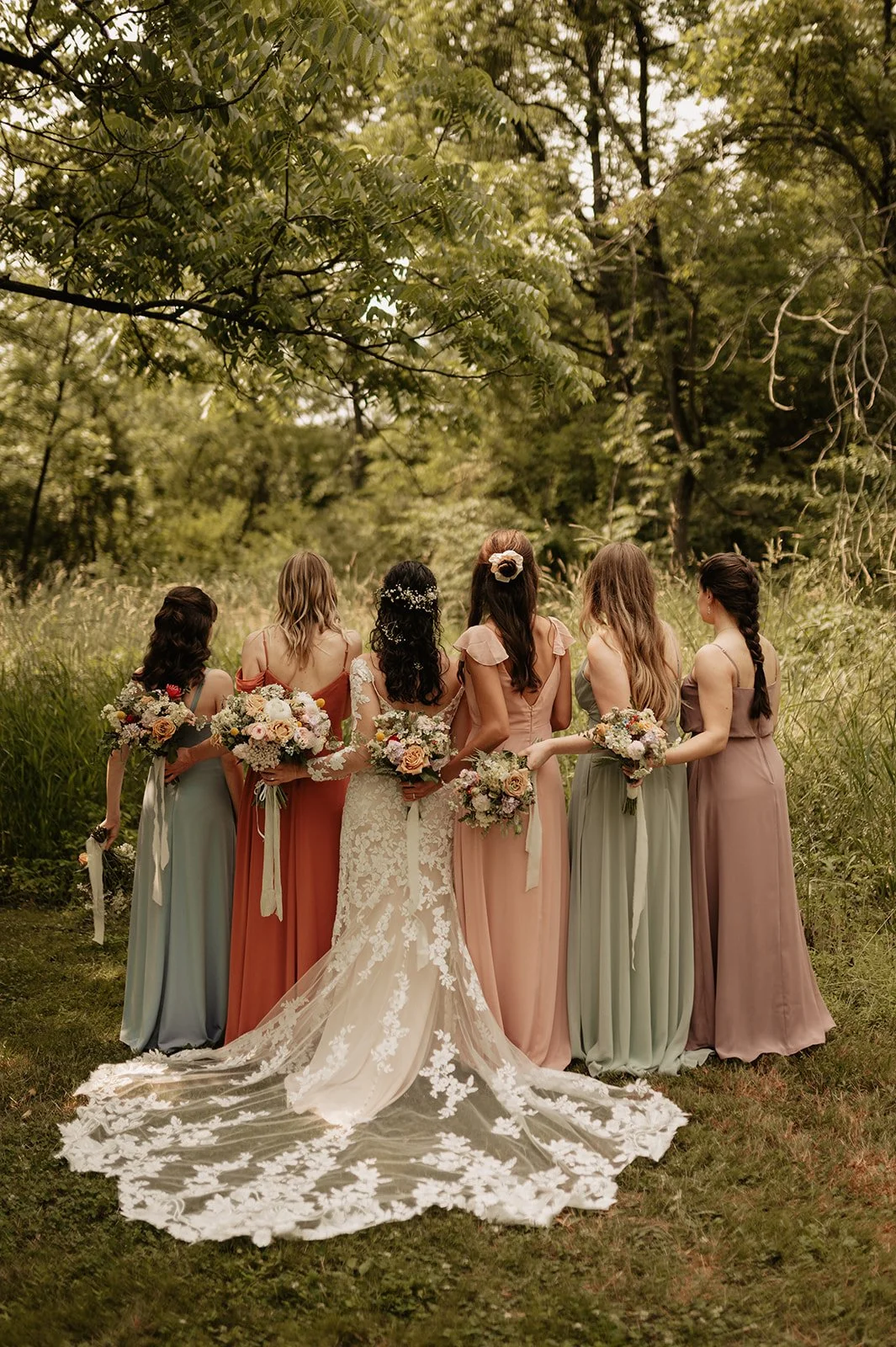 A bride in a white lace wedding gown with an extended train standing with six bridesmaids in colorful dresses, holding bouquets of flowers, in a wooded outdoor setting.