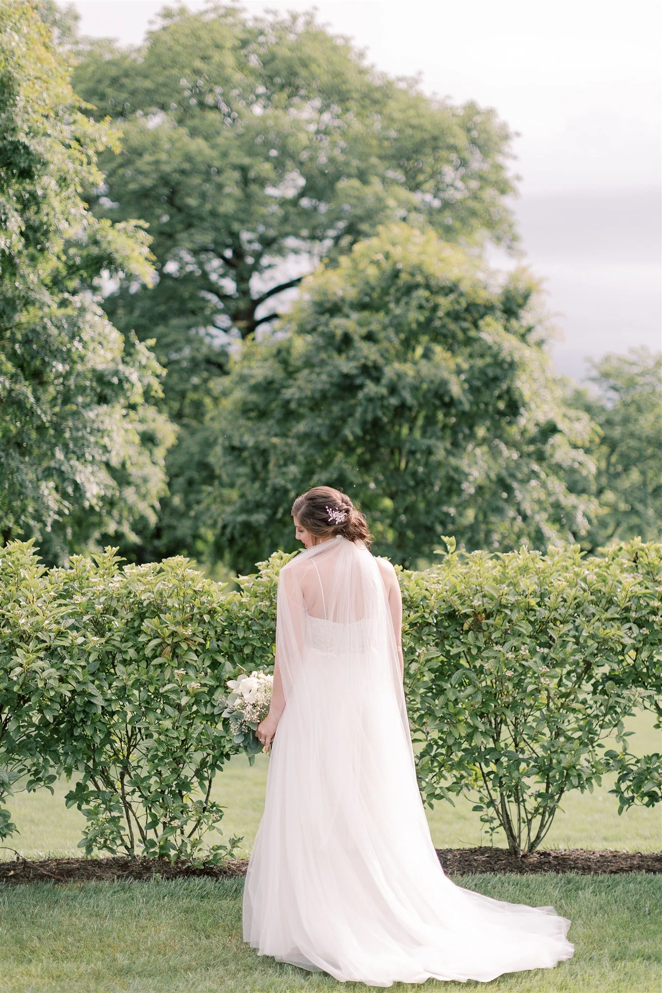 A bride standing outdoors in a garden with green bushes and trees, holding a bouquet of white flowers, wearing a flowing white wedding dress with a sheer capelet and a floral hairpiece, facing downward.