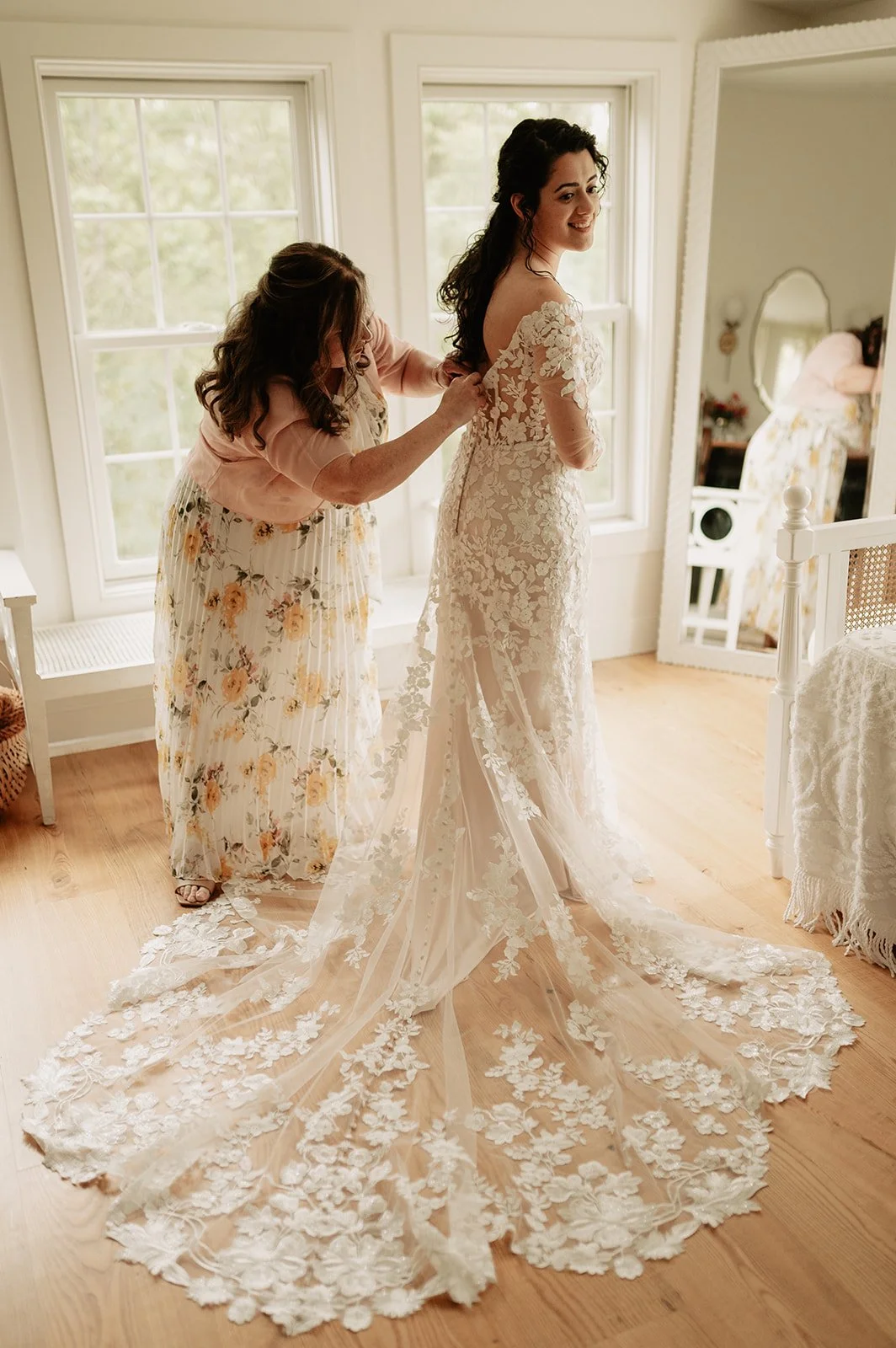 A bride getting help with her wedding dress from a woman, possibly her mother, in a well-lit room with large windows. The bride is smiling and wearing a lace wedding gown with a long train. The room has a wooden floor, a mirror, and a table covered w