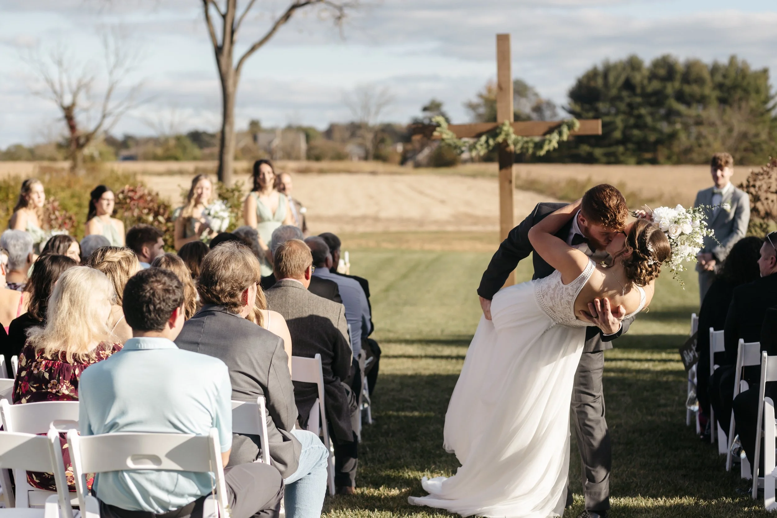 A bride and groom share a kiss during their outdoor wedding ceremony, with guests seated in front, under a wooden cross decorated with flowers, in a scenic rural landscape.