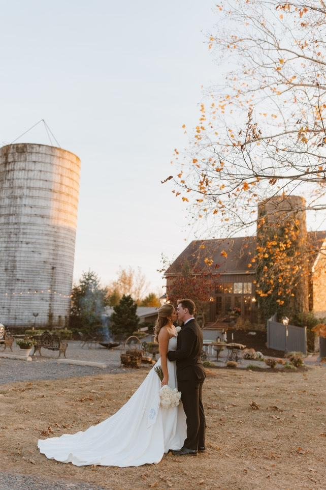 A bride and groom standing close together outdoors during sunset, with a rustic barn and silo in the background, surrounded by autumn trees and outdoor decorations.