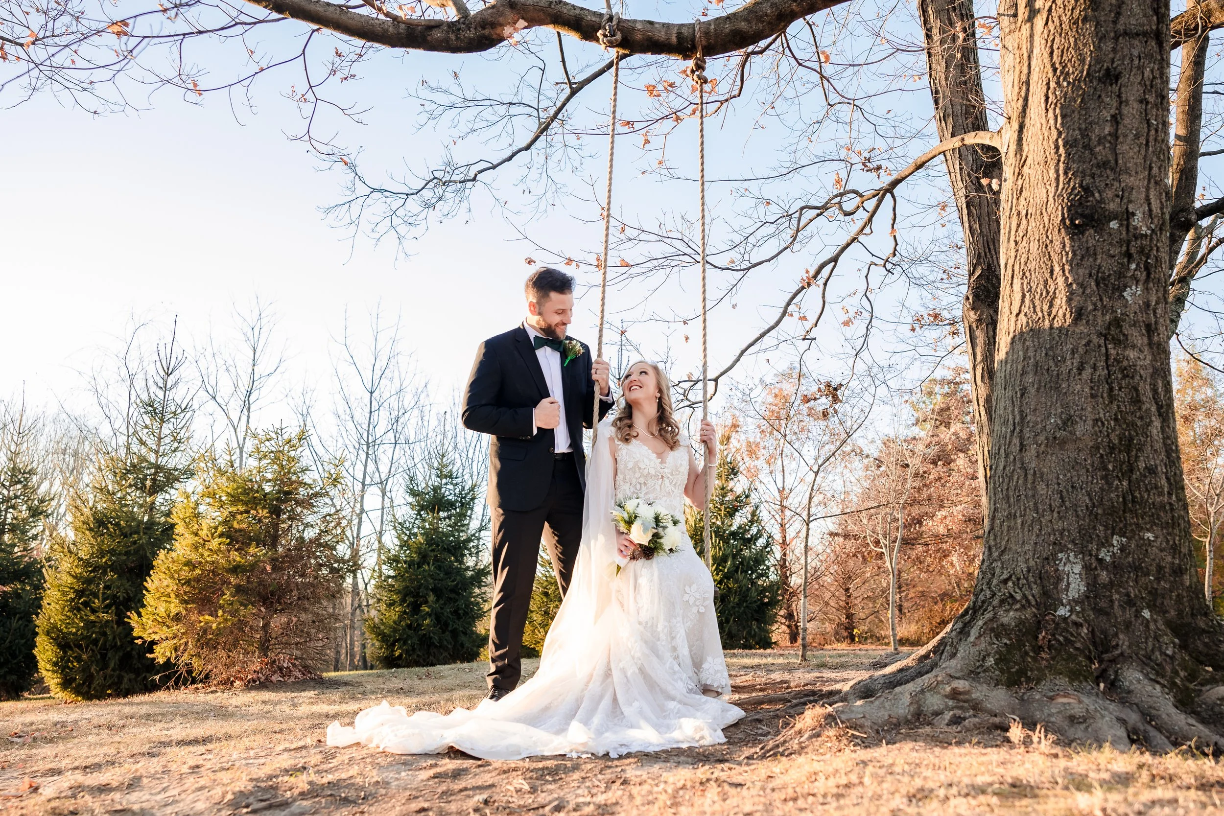 A bride and groom standing under a large tree in an outdoor setting during fall, with the bride holding a bouquet and smiling at the groom, who is wearing a black suit and bow tie, smiling back at her.
