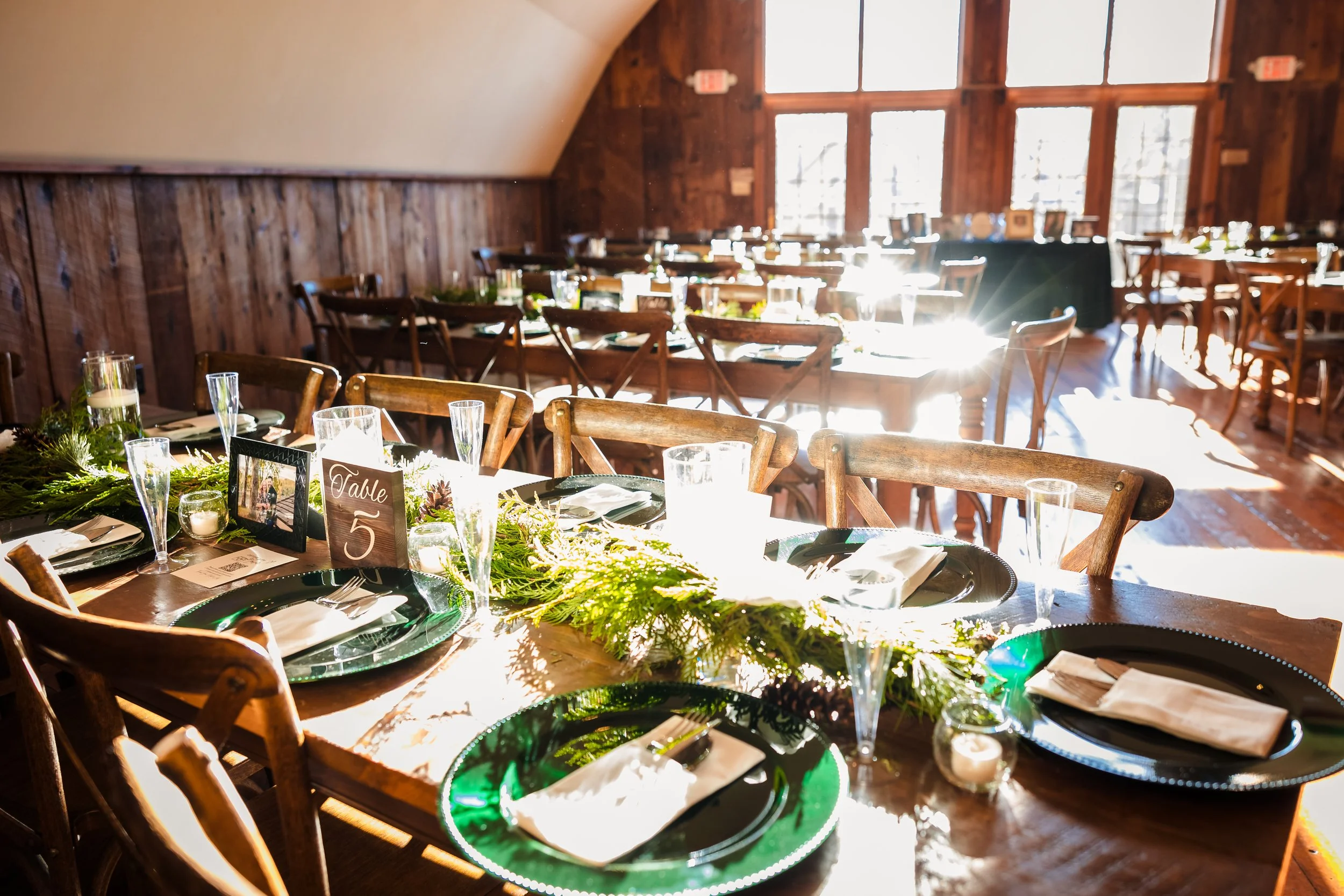 A room decorated for a celebration with a table set with green and white decorations, candles, and table number signs, illuminated by sunlight coming through large windows, with wooden chairs surrounding the table.