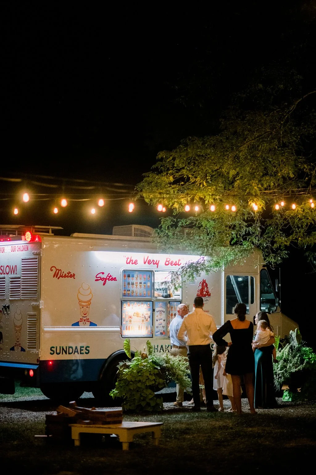Nighttime scene of a brightly lit ice cream truck with people gathered around, illuminated by string lights overhead.