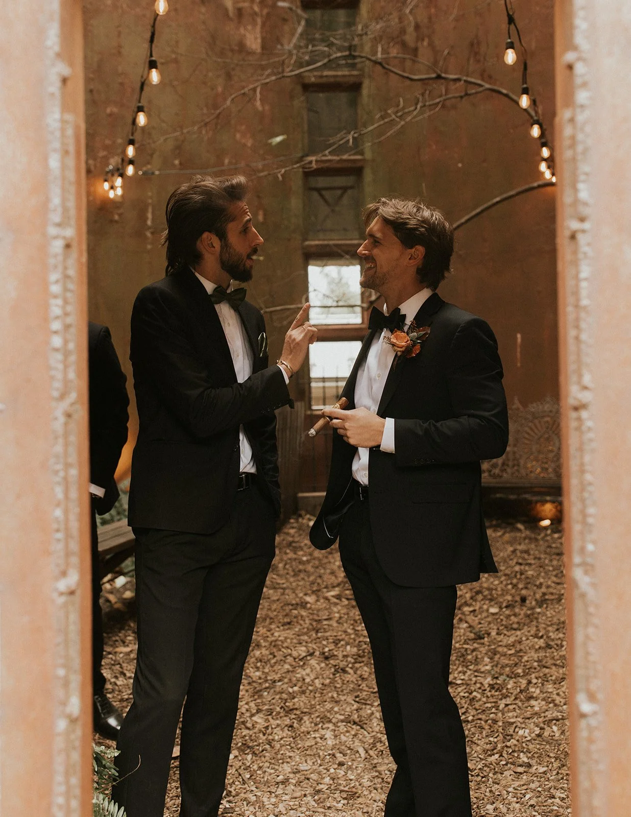 Two men in tuxedos talking and smiling at each other at a wedding or formal event, surrounded by string lights and rustic decor.