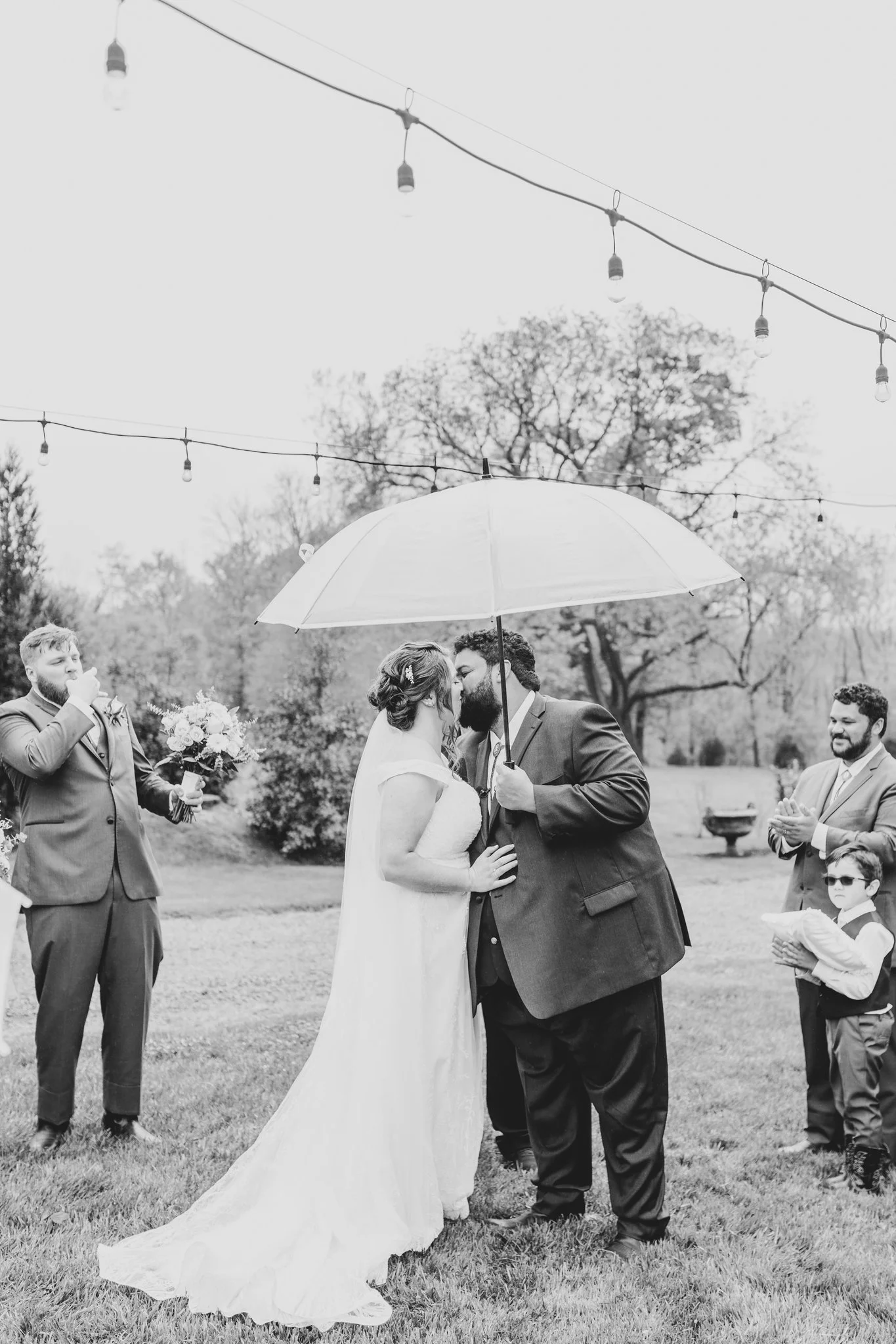 A black and white photo of a wedding ceremony outdoors, with the bride and groom kissing Under an umbrella. The bride wears a wedding dress, and the groom wears a suit. Guests, including a man in a suit holding a bouquet and a young boy in glasses, s