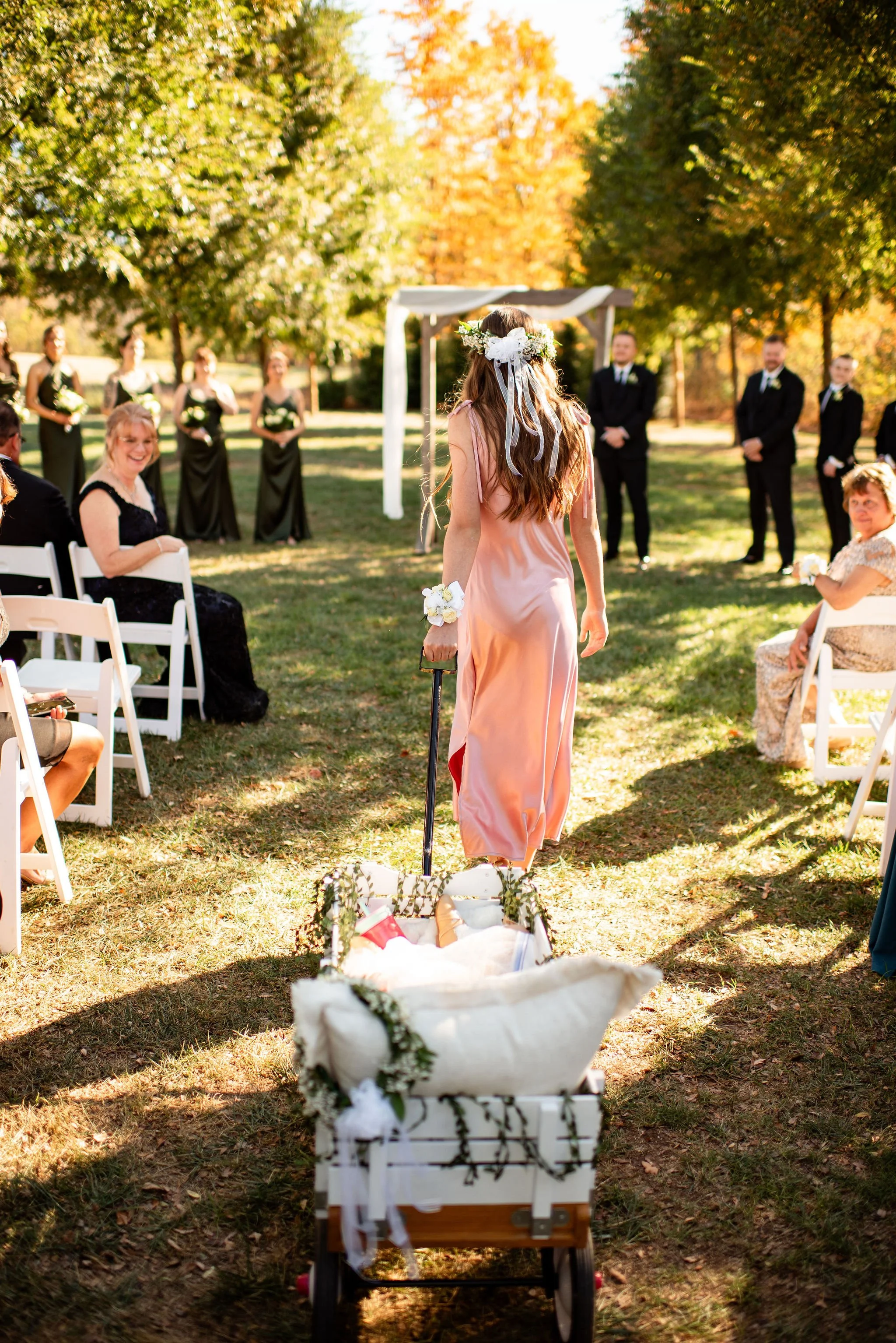 A woman in a pink satin dress with a floral headband walks towards an outdoor wedding ceremony, pushing a white cart decorated with flowers. Guests are seated on white chairs, and a group of men and women dressed in formal black attire stand near a s