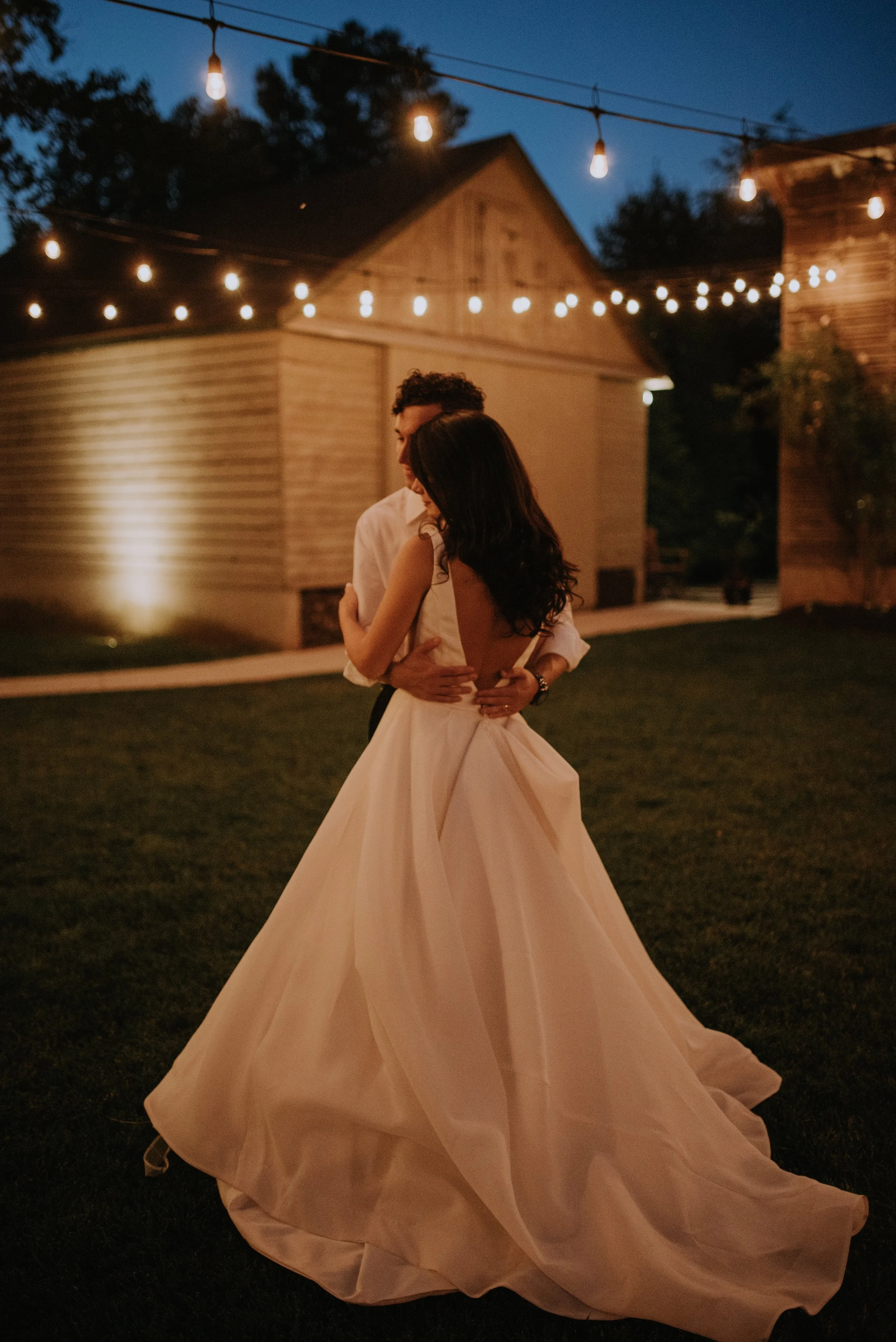 A couple dancing outdoors at night under string lights with a barn in the background. The woman is wearing a long cream-colored dress, and the man is dressed in a light shirt.