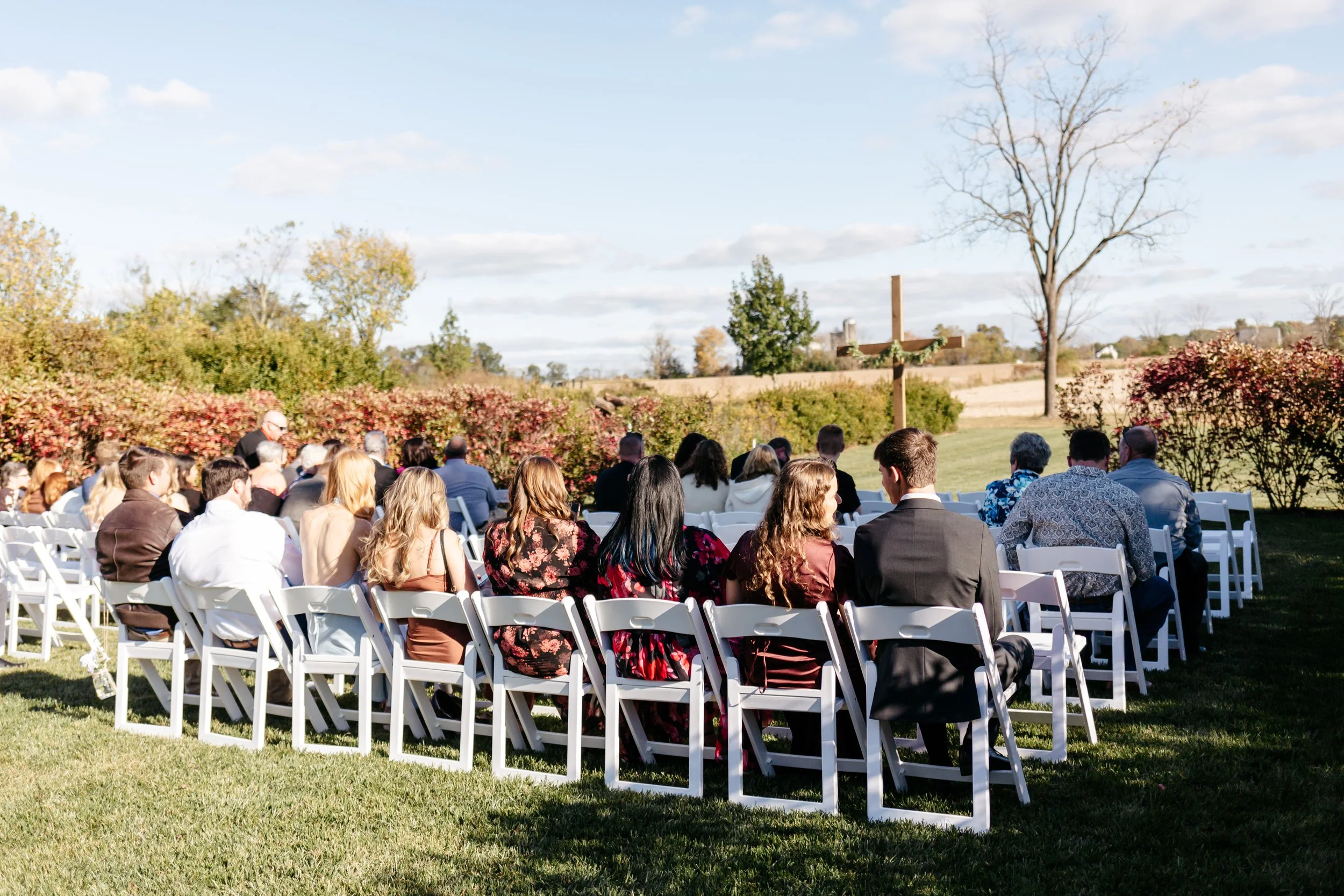 A gathering of people seated on white folding chairs outdoors on a sunny day, with a cross and trees in the background, suggesting a religious or memorial ceremony.