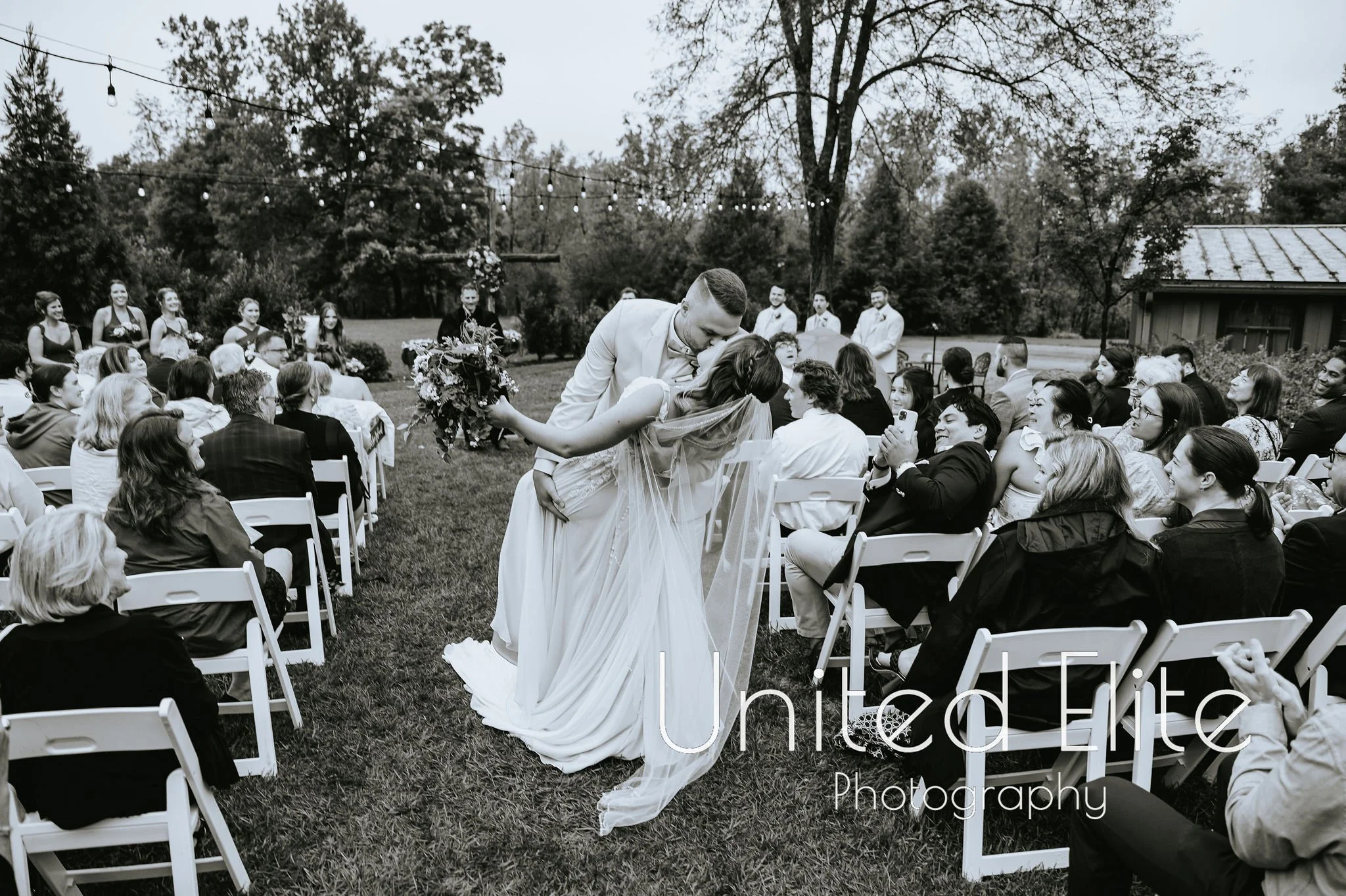 A couple sharing a kiss during their outdoor wedding ceremony surrounded by seated guests and bridesmaids, with trees and string lights in the background.