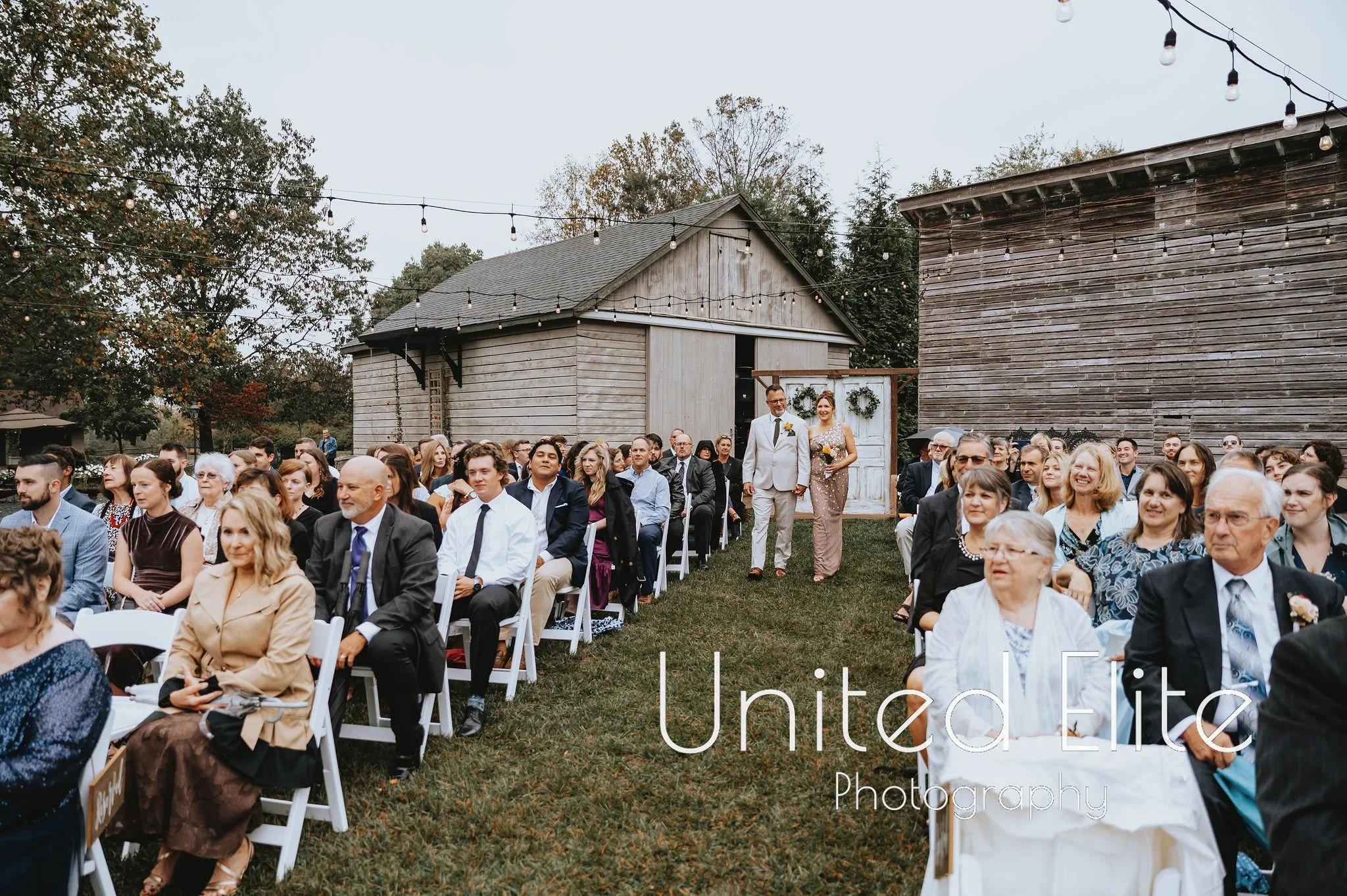 A large group of wedding guests seated outdoors, with a couple walking down the aisle in front of rustic barn buildings decorated with greenery and wreaths, and string lights overhead.