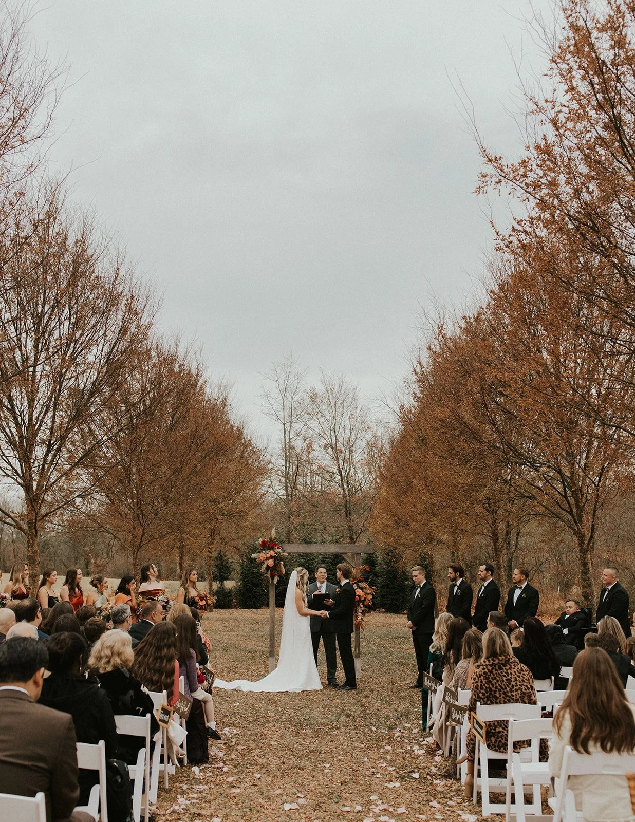 Outdoor wedding ceremony with bride and groom exchanging vows under a decorated arch, surrounded by wedding party and guests, in a park with trees showing autumn colors.