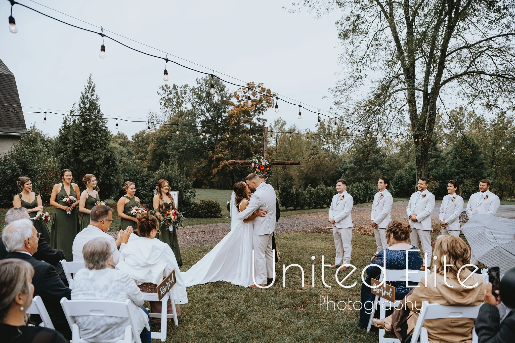 A wedding ceremony outdoors with a bride and groom kissing, surrounded by bridesmaids and groomsmen, with guests seated on white chairs under string lights and trees.