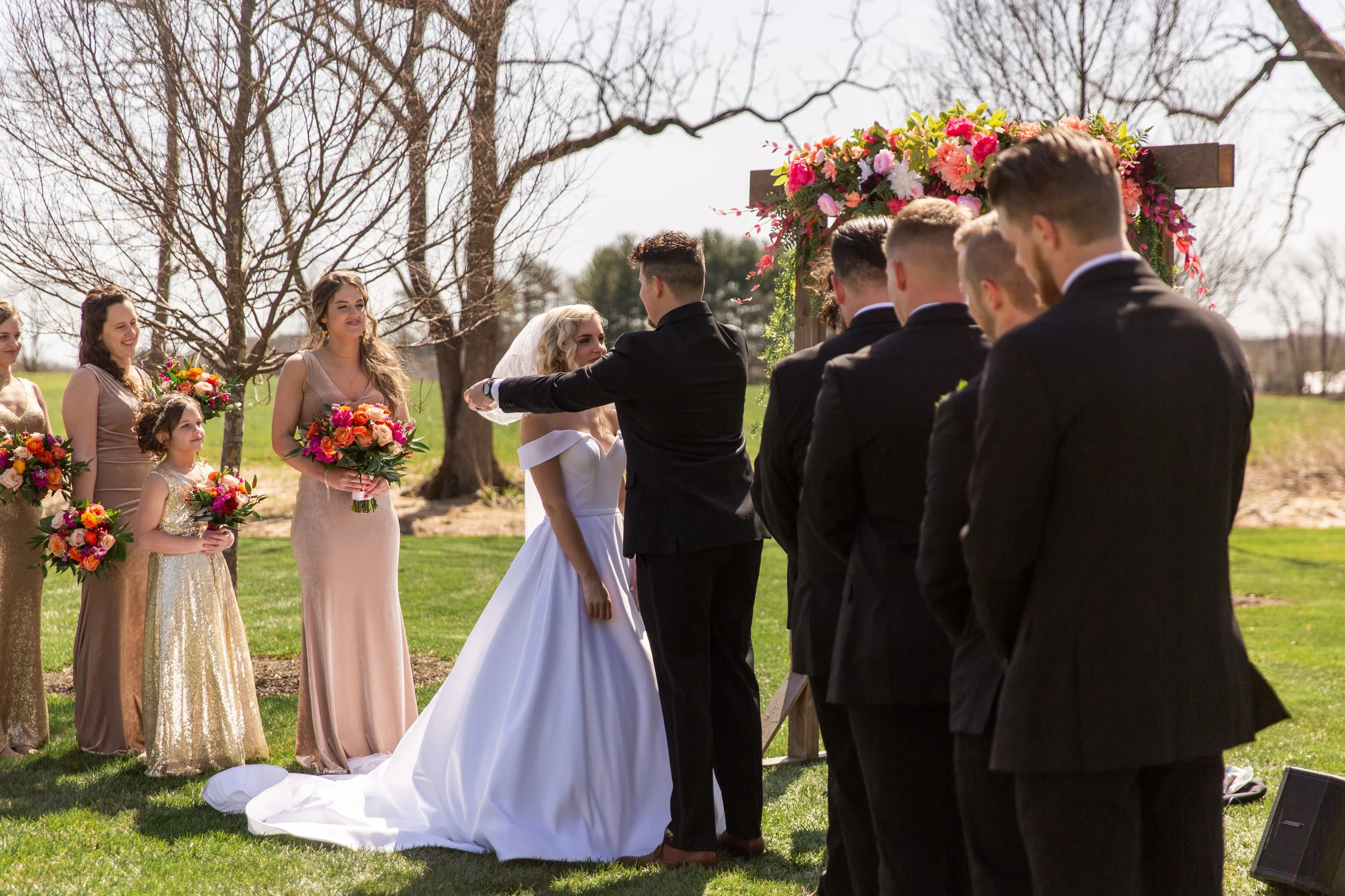 An outdoor wedding ceremony with the bride, groom, bridesmaids, and groomsmen standing under a floral arch on a grass lawn with leafless trees in the background.
