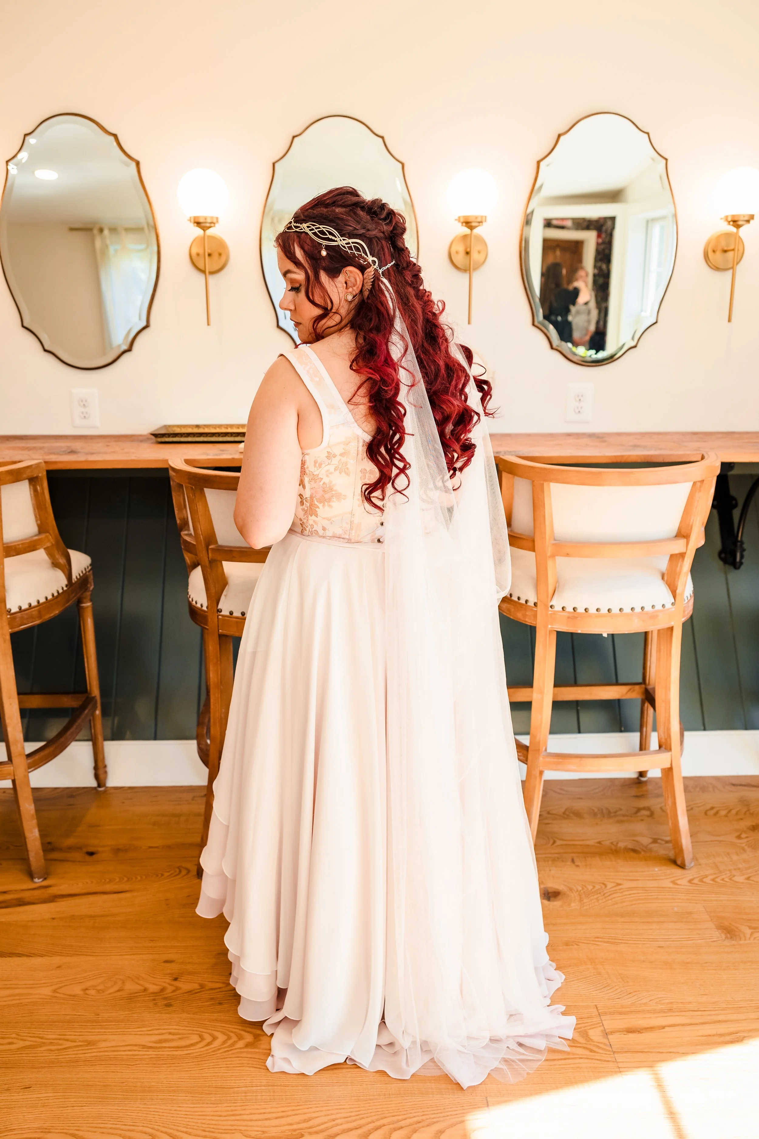 A bride with long, curled red hair, wearing a floral lace wedding dress and a headpiece with a veil, standing indoors in front of a mirror with three decorative mirrors and wall-mounted lamps, wooden chairs, and a wooden floor.