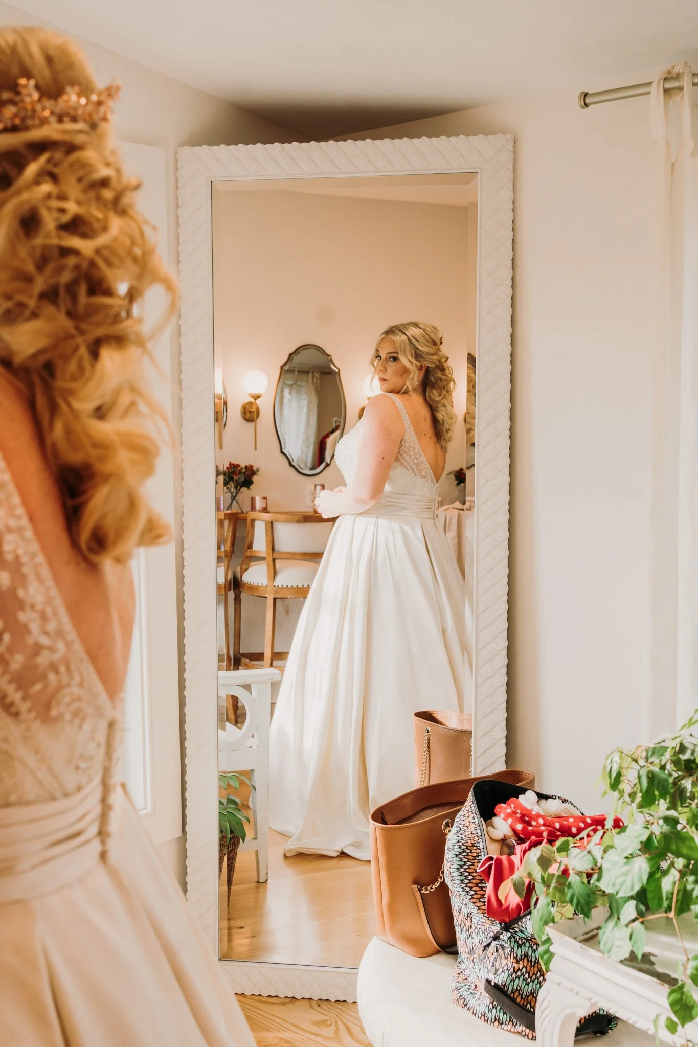 A bride in a white wedding gown looking at herself in a full-length mirror in a softly lit room decorated with flowers and furniture, with a visible dresser in the background.
