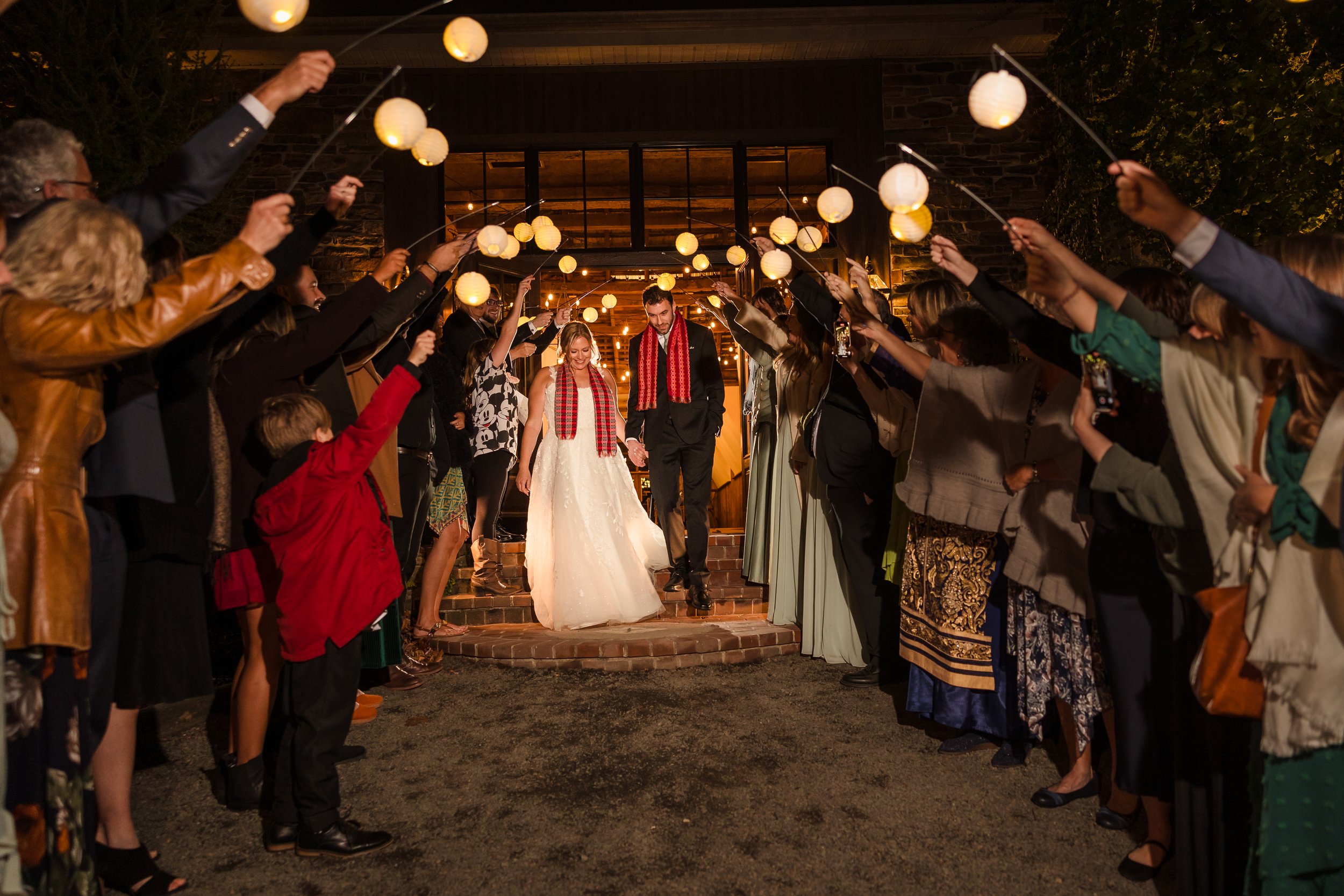 Bride and groom walking down stairs at night surrounded by friends and family holding paper lanterns and taking pictures.