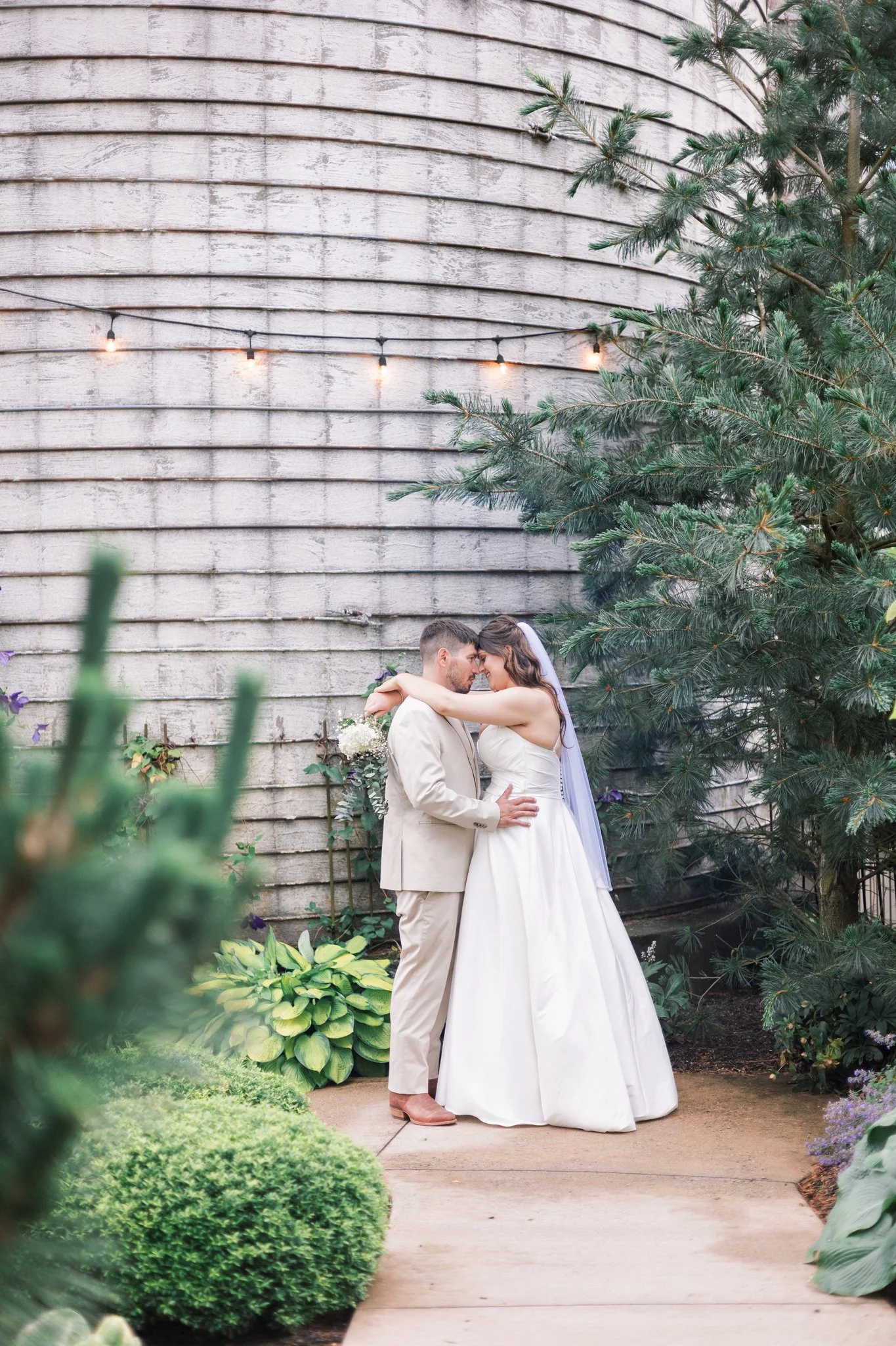 A bride and groom embracing and touching foreheads in a garden with greenery, flowers, and an old wooden wall in the background, decorated with string lights, during their wedding.