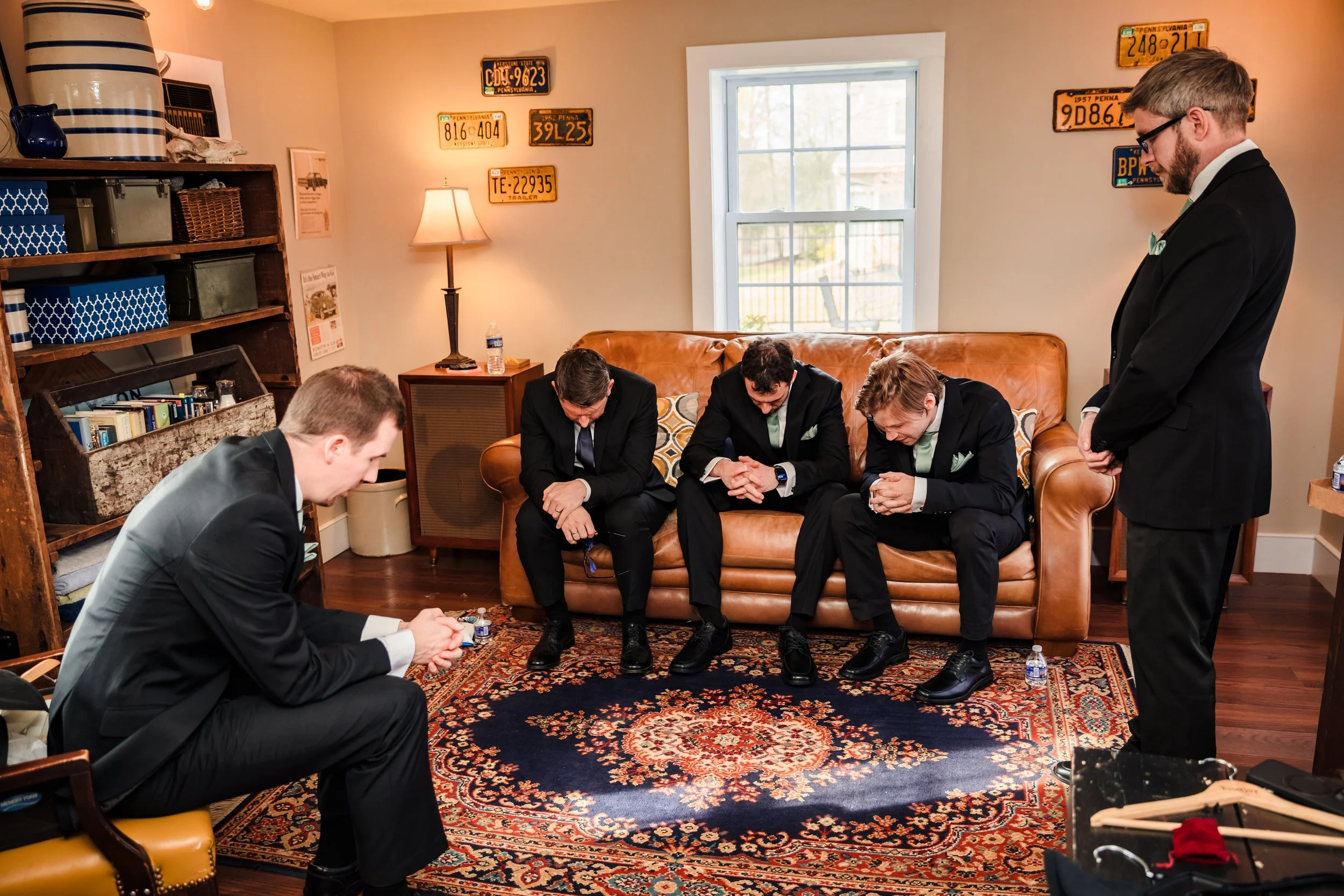 Five men dressed in suits are kneeling and bowing their heads in a prayer or moment of silence in a living room. One man is standing on the right side of the image, also with his head bowed. The room has a window, a bookshelf, and various license plates on the walls.
