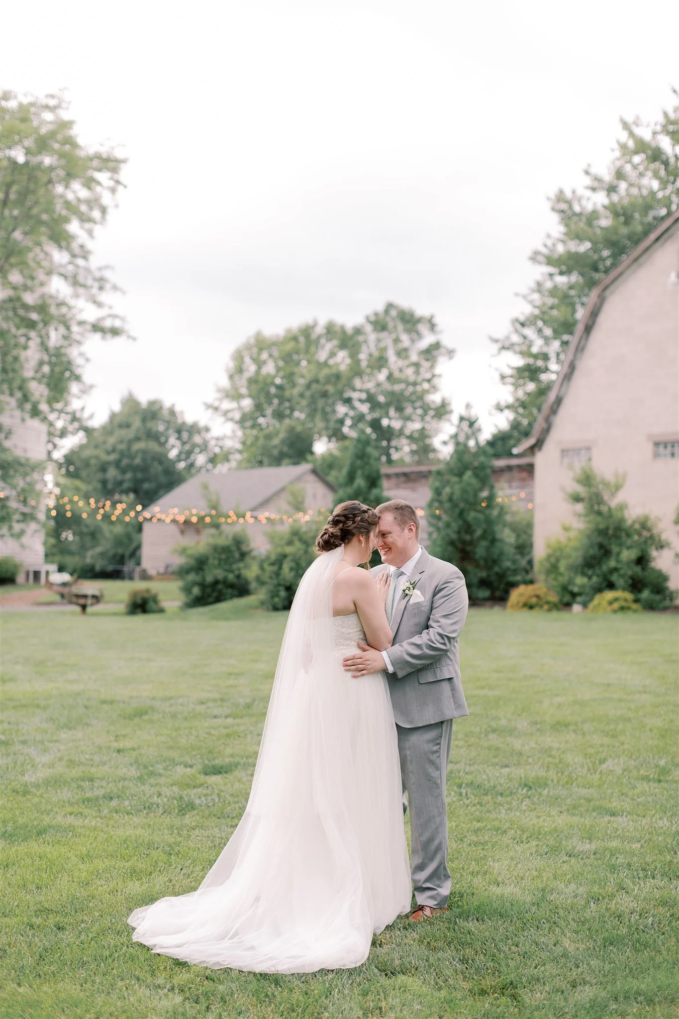 A bride and groom are standing close together outdoors on a grassy area, sharing an intimate moment, with a background of houses, trees, and string lights.