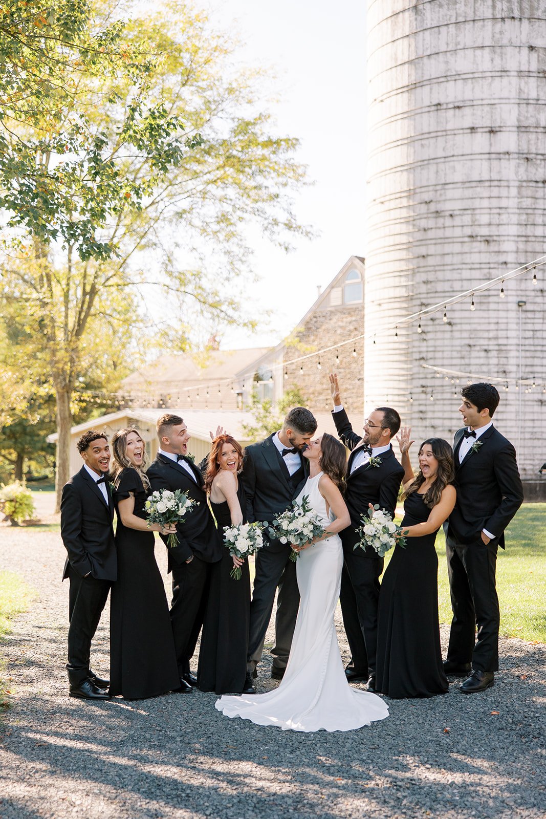 A group of happily dressed wedding guests, including bride and groom, celebrating outdoors on a sunny day with trees and a large barn or silo in the background.
