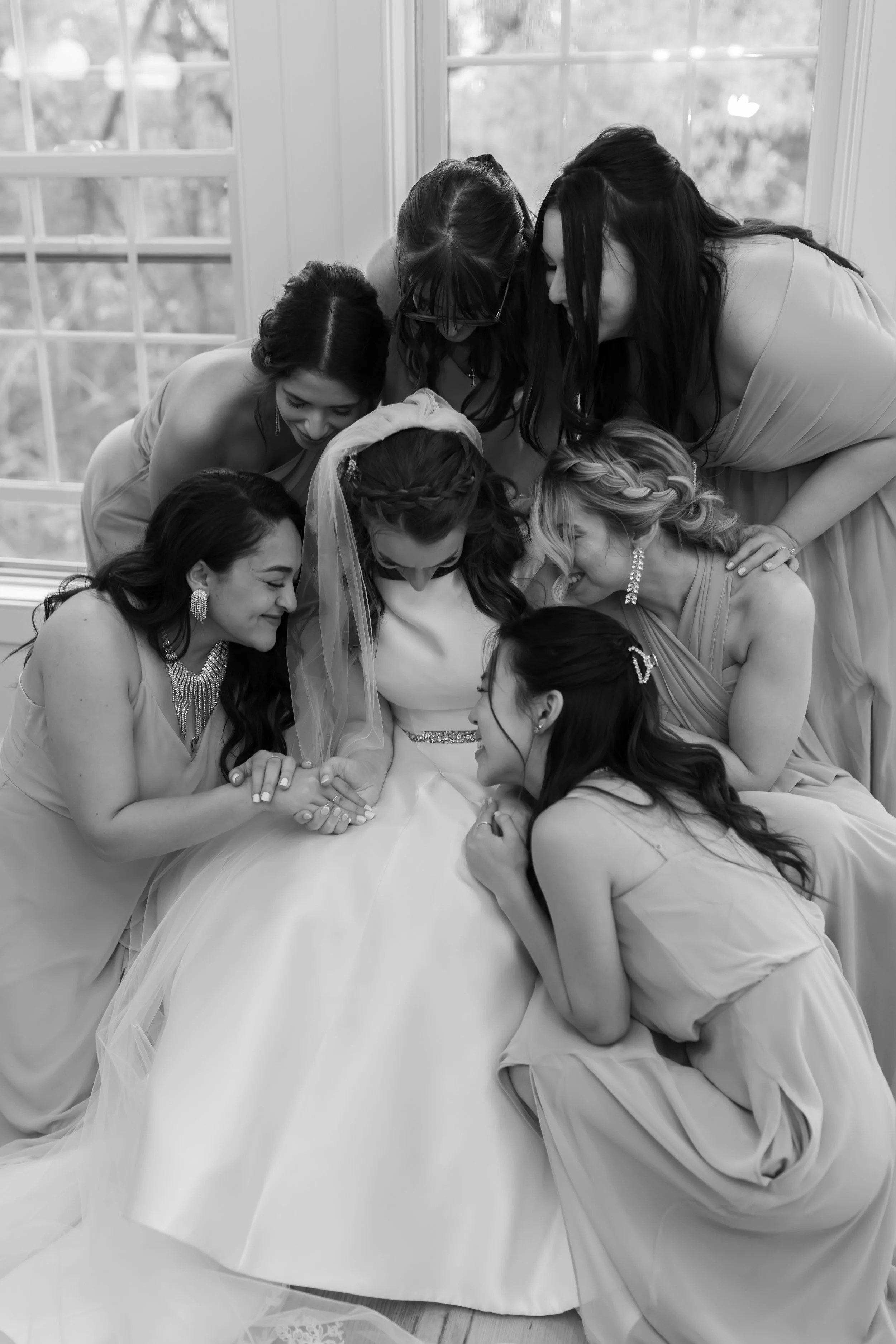 Black and white photo of a bride surrounded by her bridesmaids, all leaning over and looking at the bride's hand, which is being held by a bridesmaid. The women are inside near a large window with trees visible outside.