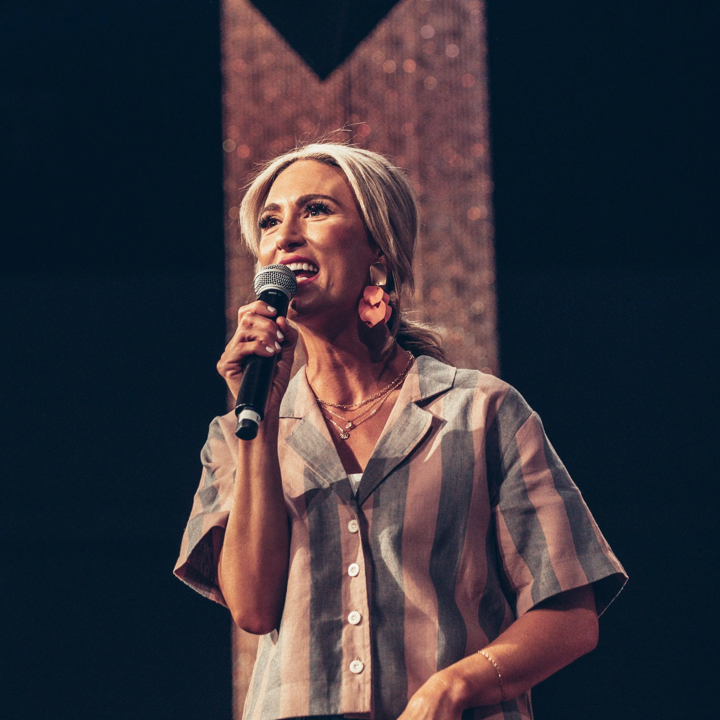 A woman with blonde hair smiling and holding a microphone, wearing a blazer and large earrings, on stage with a dark background and a blurry, illuminated graphic behind her.
