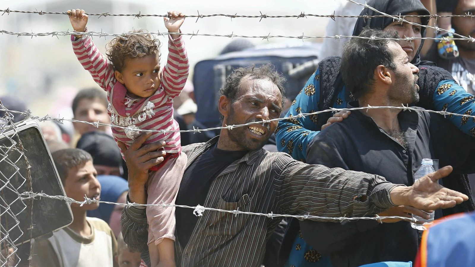 A Syrian refugee carries his child as he waits behind border fences to cross into Turkey. Cover credit: WordPress