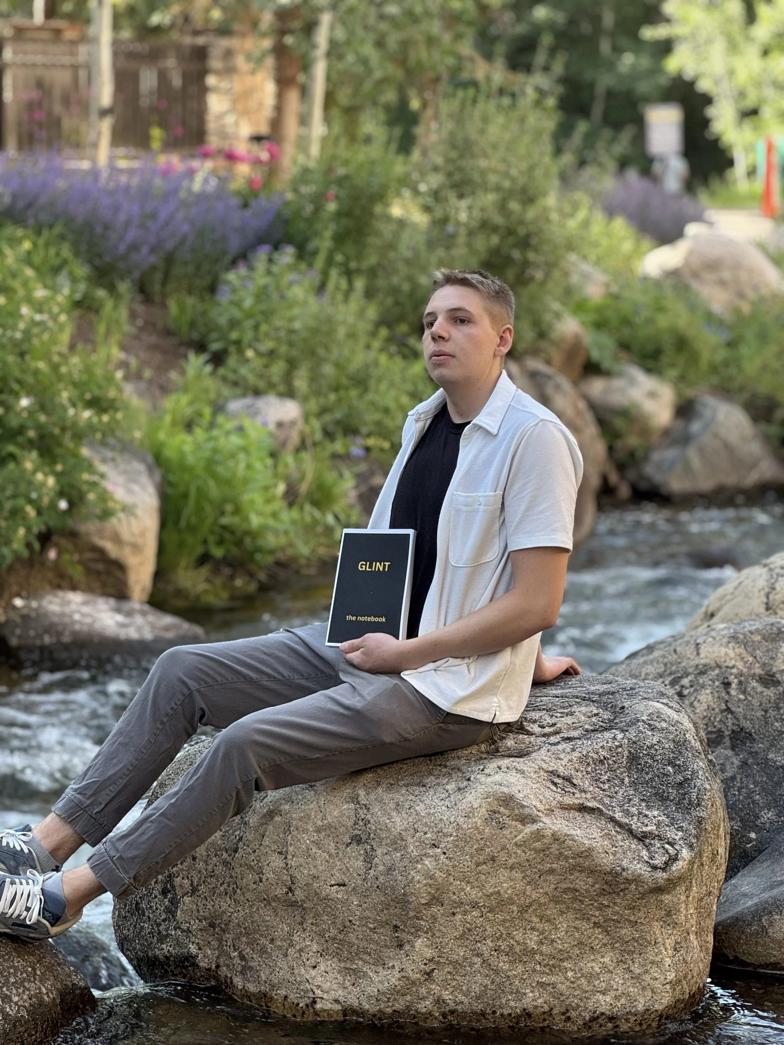 A young man sitting on a large rock in a stream, holding a black notebook with yellow text titled 'GLINT' and 'the notebook', surrounded by greenery and rocks.
