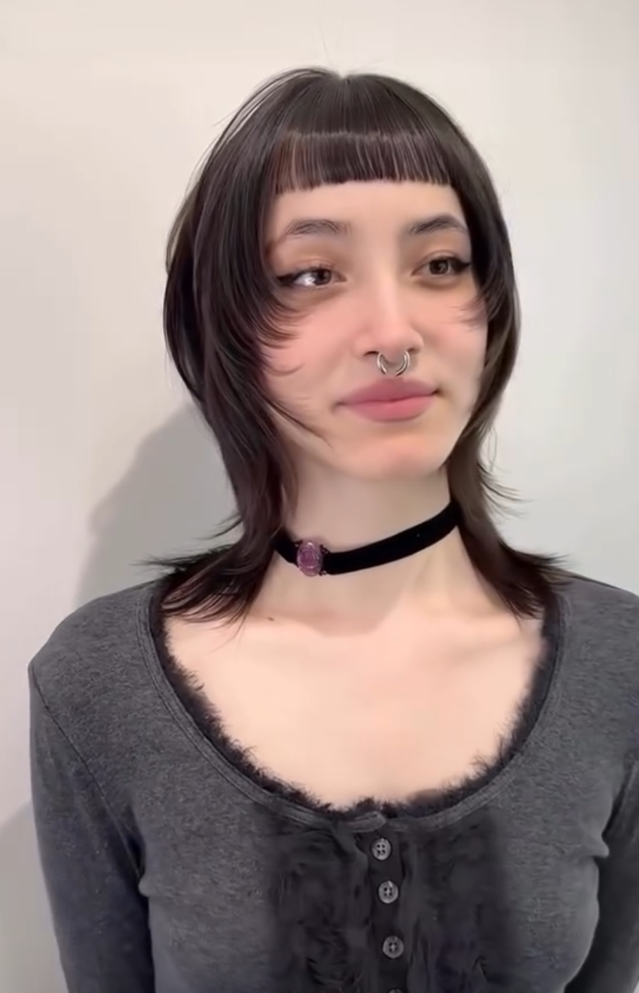 A young woman with short, layered brown hair and blunt bangs, standing against a plain, light-colored wall. She has a septum piercing, is wearing a black choker with a purple gemstone, and a gray long-sleeve top with a buttoned neckline.
