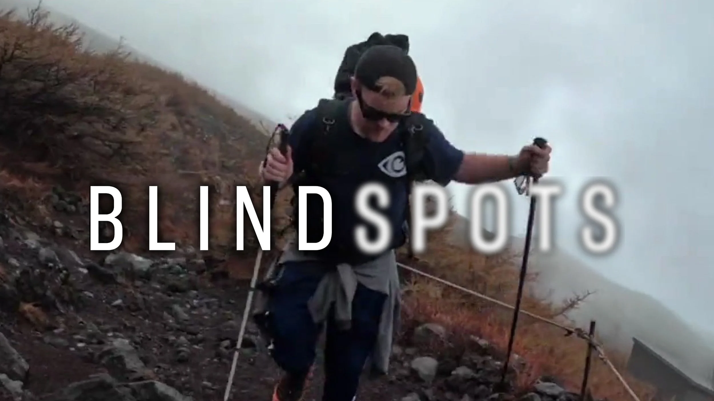 James Rath using his White Cane and a trekking pole, climbs up Mount Fuji. The Blindspots logo is overlaid. Spots is slightly blurred.