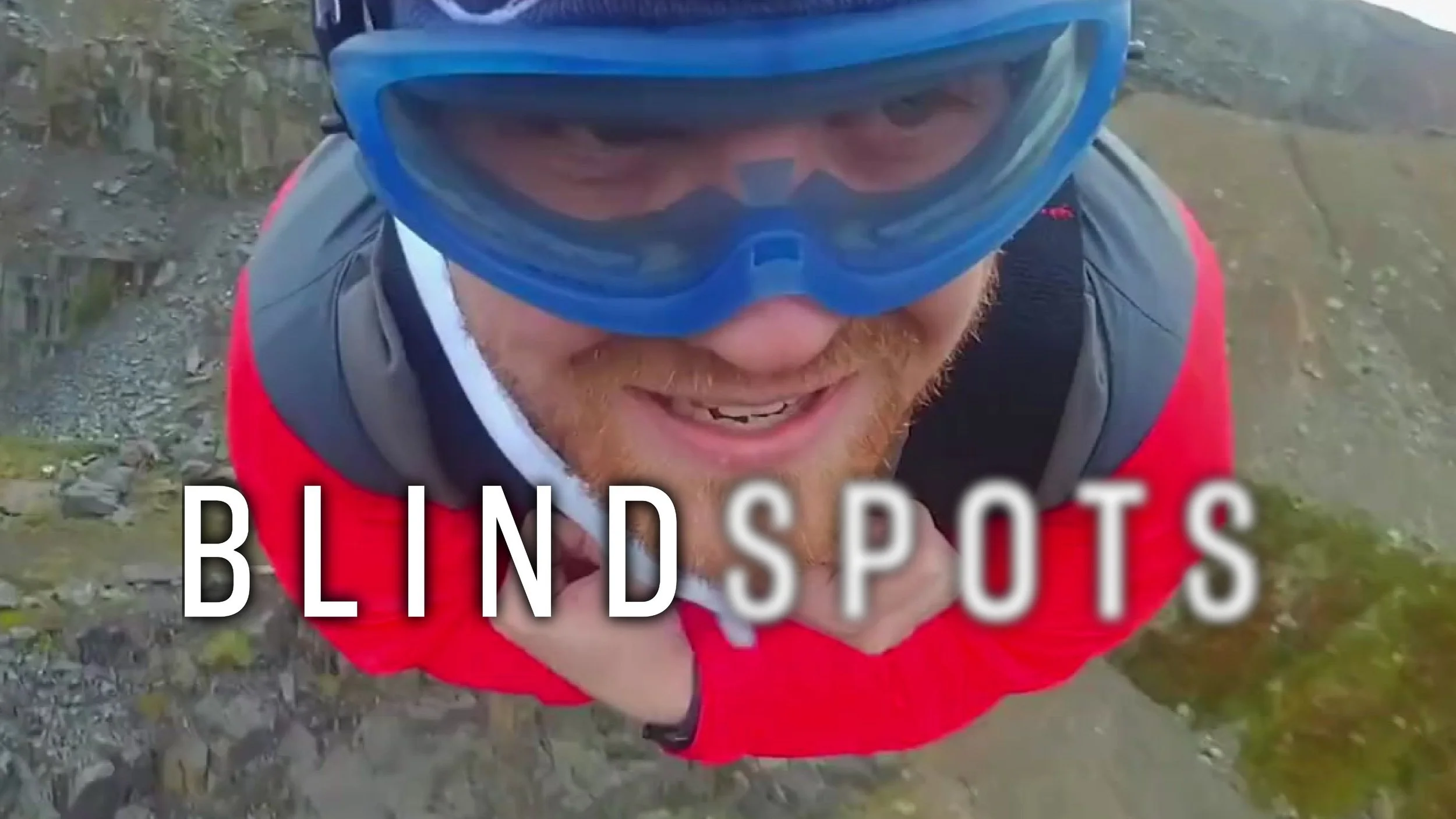 James Rath zip lines through rocky mountains wearing a blue eye mask and clenching his teeth. The Blindspots logo is overlaid. Spots is slightly blurred.