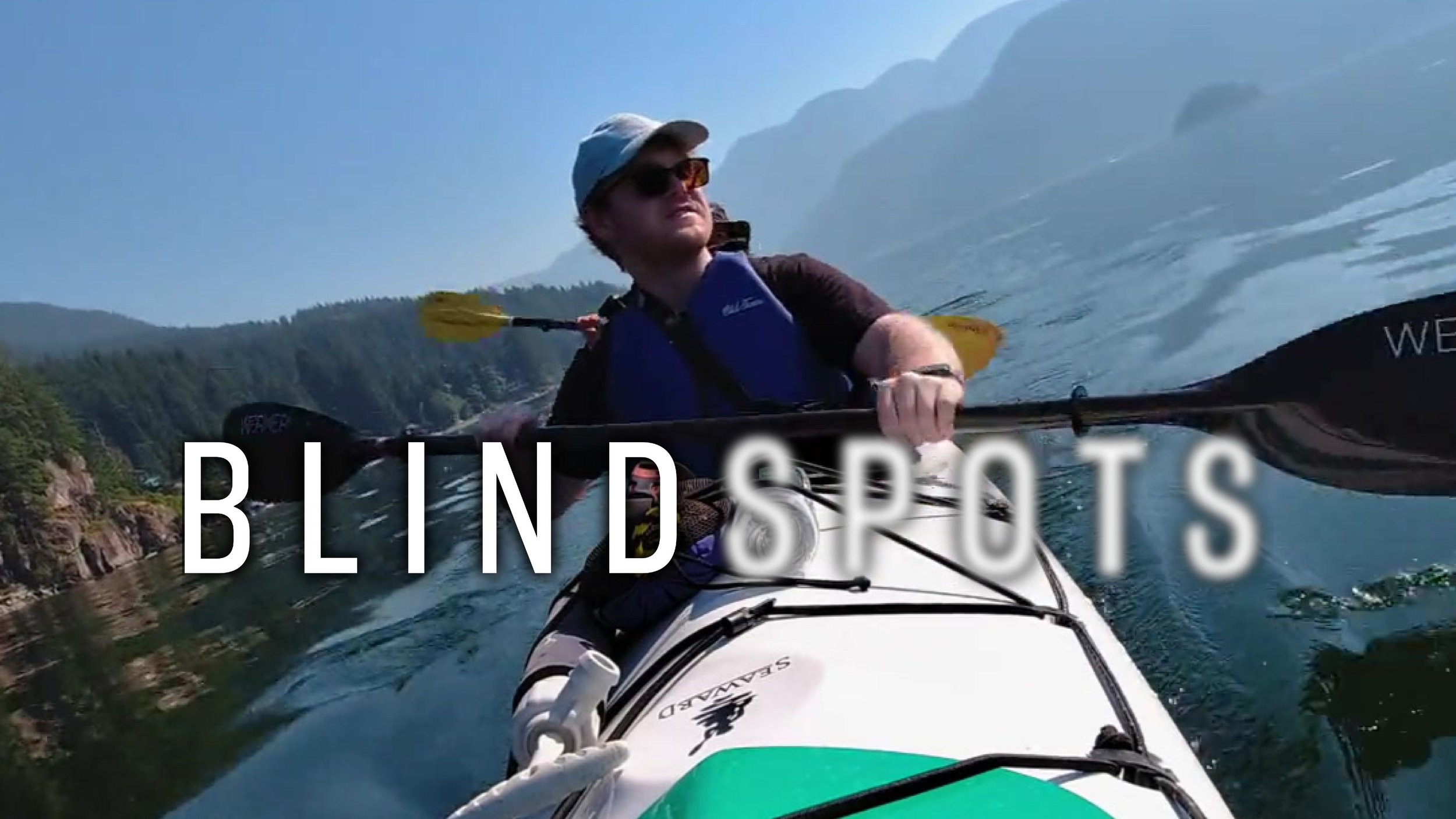 James Rath along with a woman out of frame kayak on a body of water surrounded by lush mountains and woods. The Blindspots logo is overlaid. Spots is slightly blurred.
