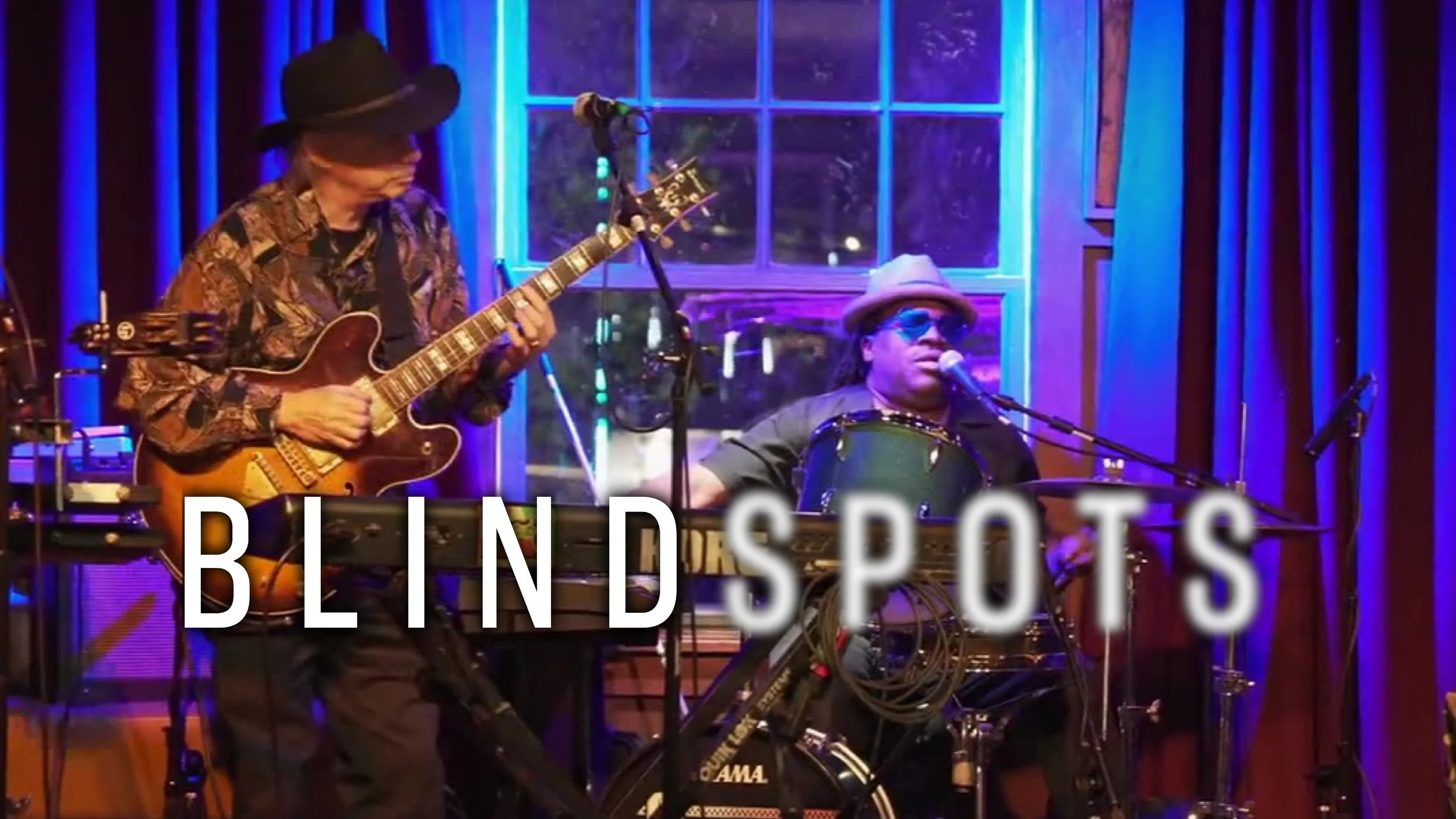 Musician, D-Madness, performs on stage with a band indoors. The Blindspots logo is overlaid. Spots is slightly blurred.