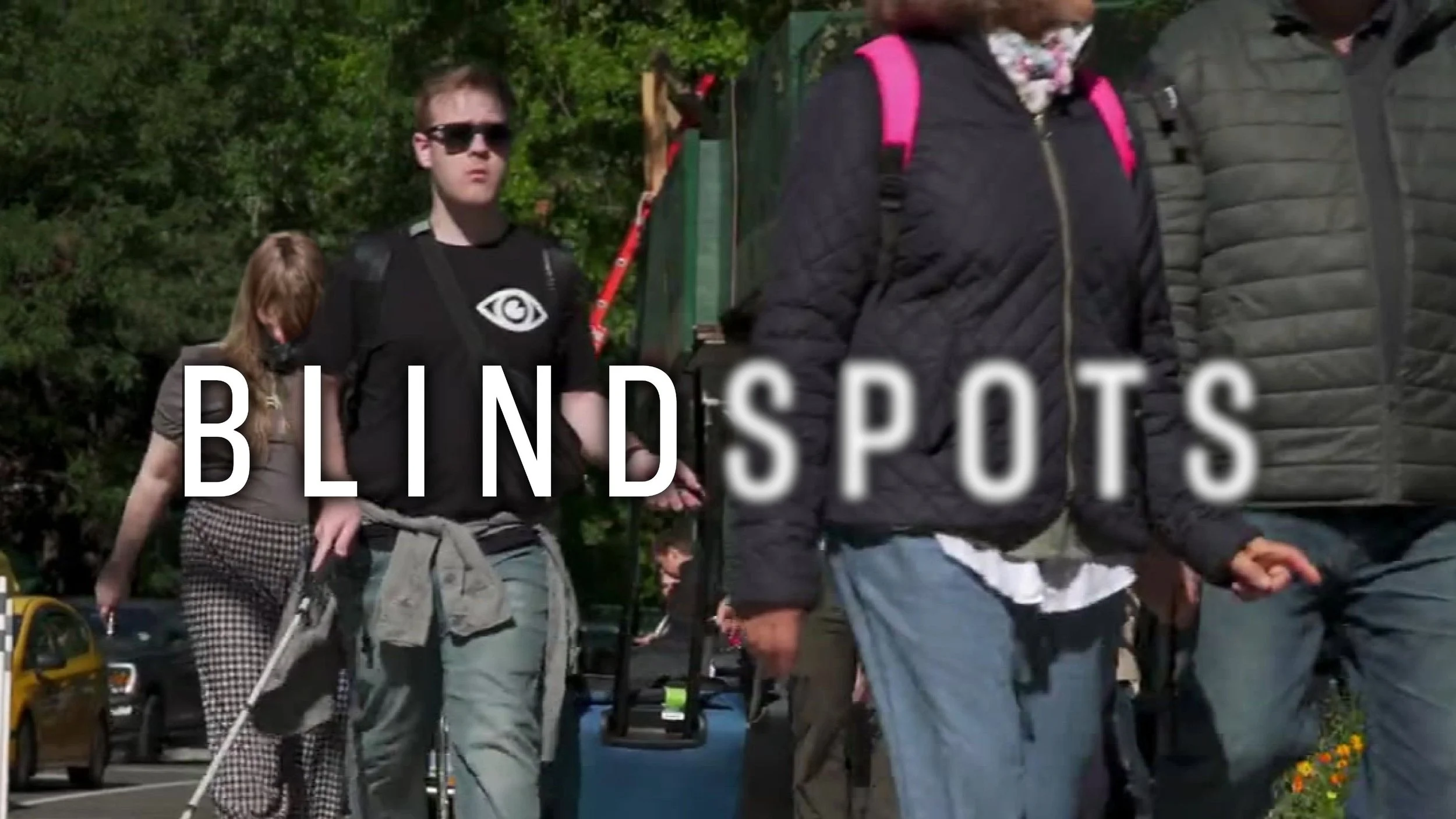 James Rath using his White Cane walks down a New York City street busy with pedestrians. The Blindspots logo is overlaid. Spots is slightly blurred.