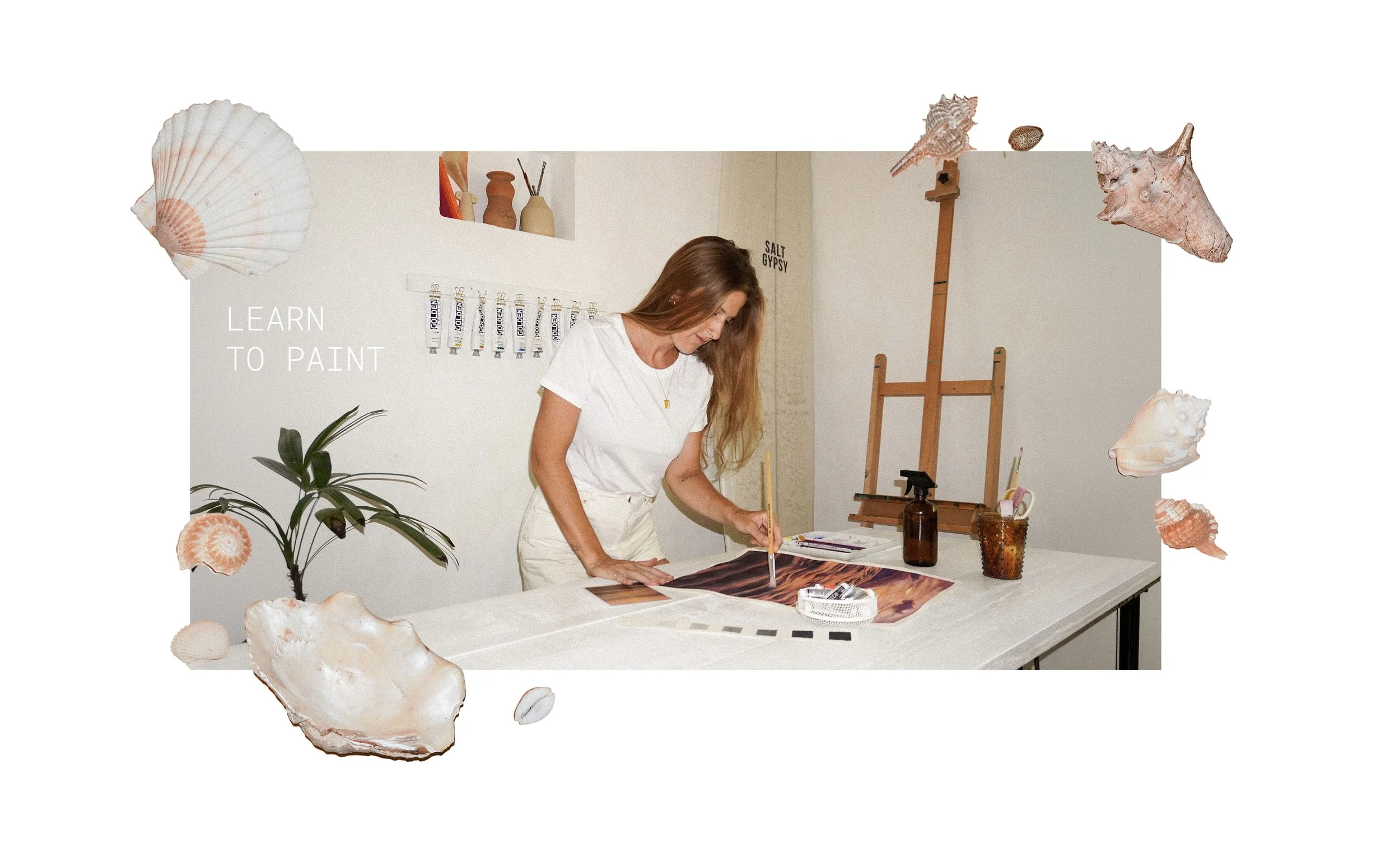 A woman with long brown hair wearing a white t-shirt and beige pants is painting on a canvas at a table in a cozy, decorated art studio.