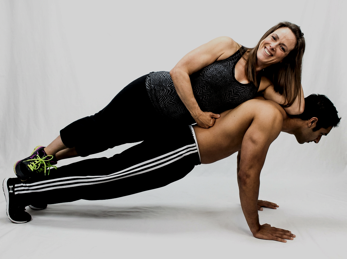 A woman with long brown hair smiling while sitting on a man's back during a work-out in a gym studio.