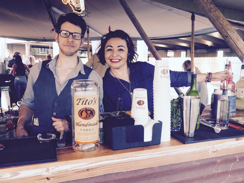 Two smiling bartenders behind a bar stocked with Tito’s