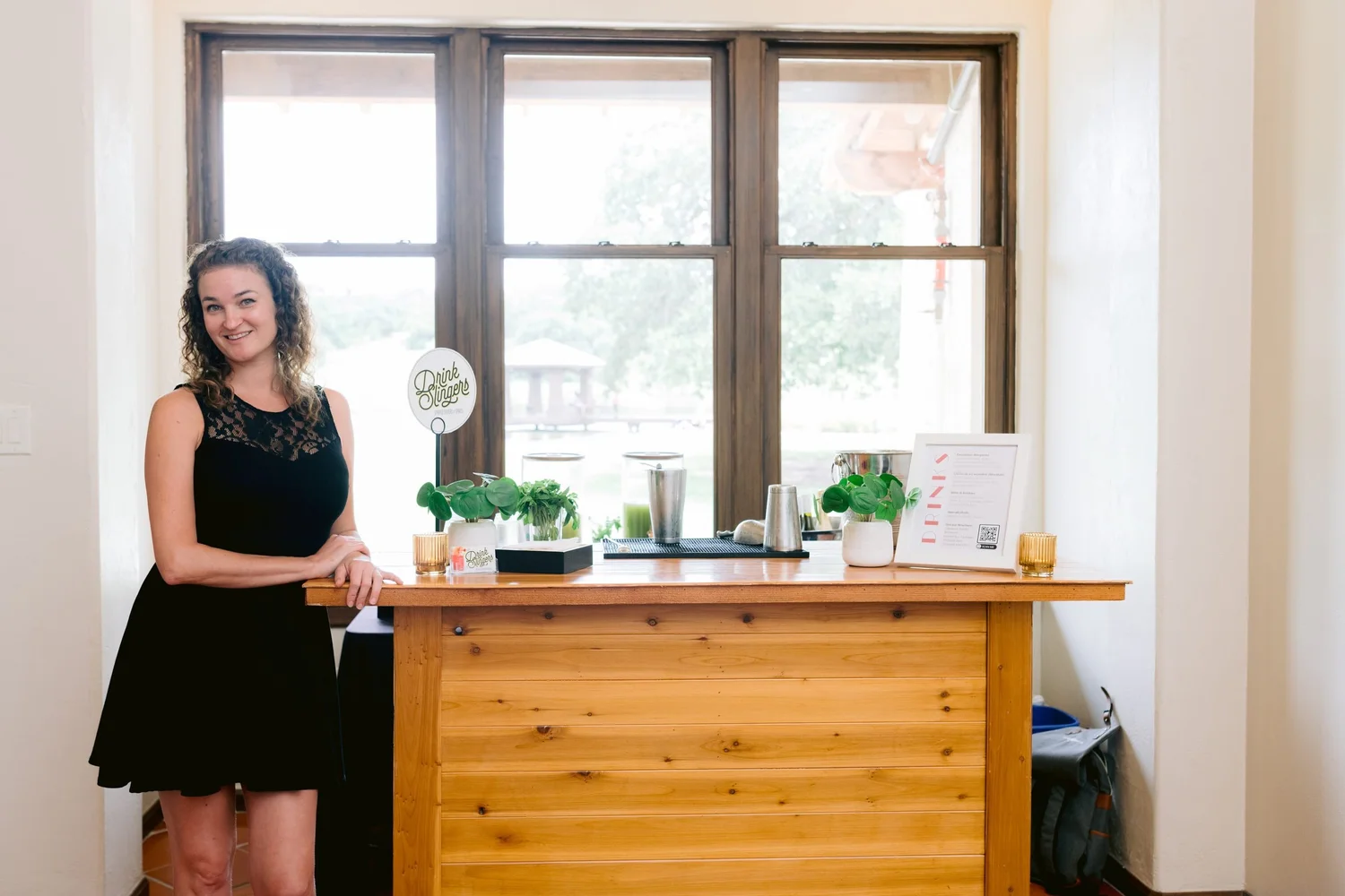 A Drink Slingers bartender beside a wooden event bar with a Drink Slingers sign, plants and a framed menu