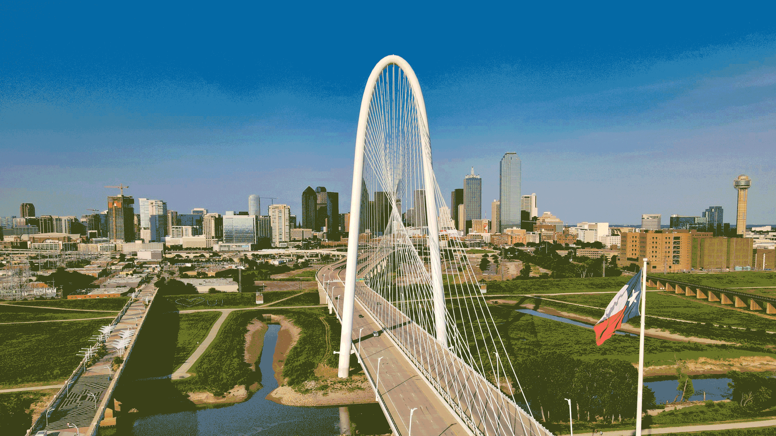 Aerial view of Dallas, Texas, featuring the Margaret Hunt Hill Bridge with a city skyline in the background, a flag, and surrounding greenery.