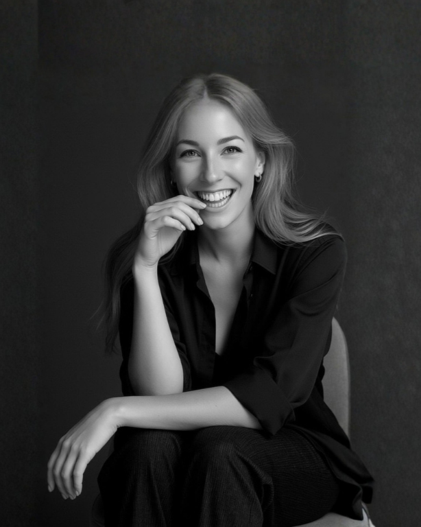 A black and white portrait of Lauren Ray, smiling with long blonde hair, wearing a dark shirt, sitting against a plain background.