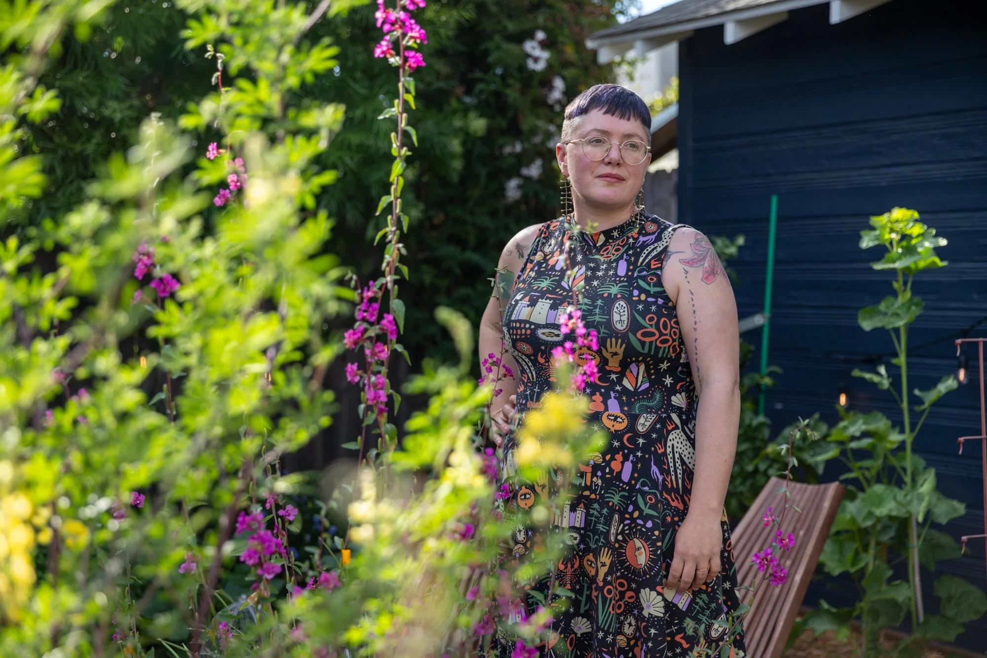 Luiza Flynn-Goodlett—a fat, short-haired, white woman wearing a mock-turtleneck dress patterned with graphics in a tarot—stands in a garden.