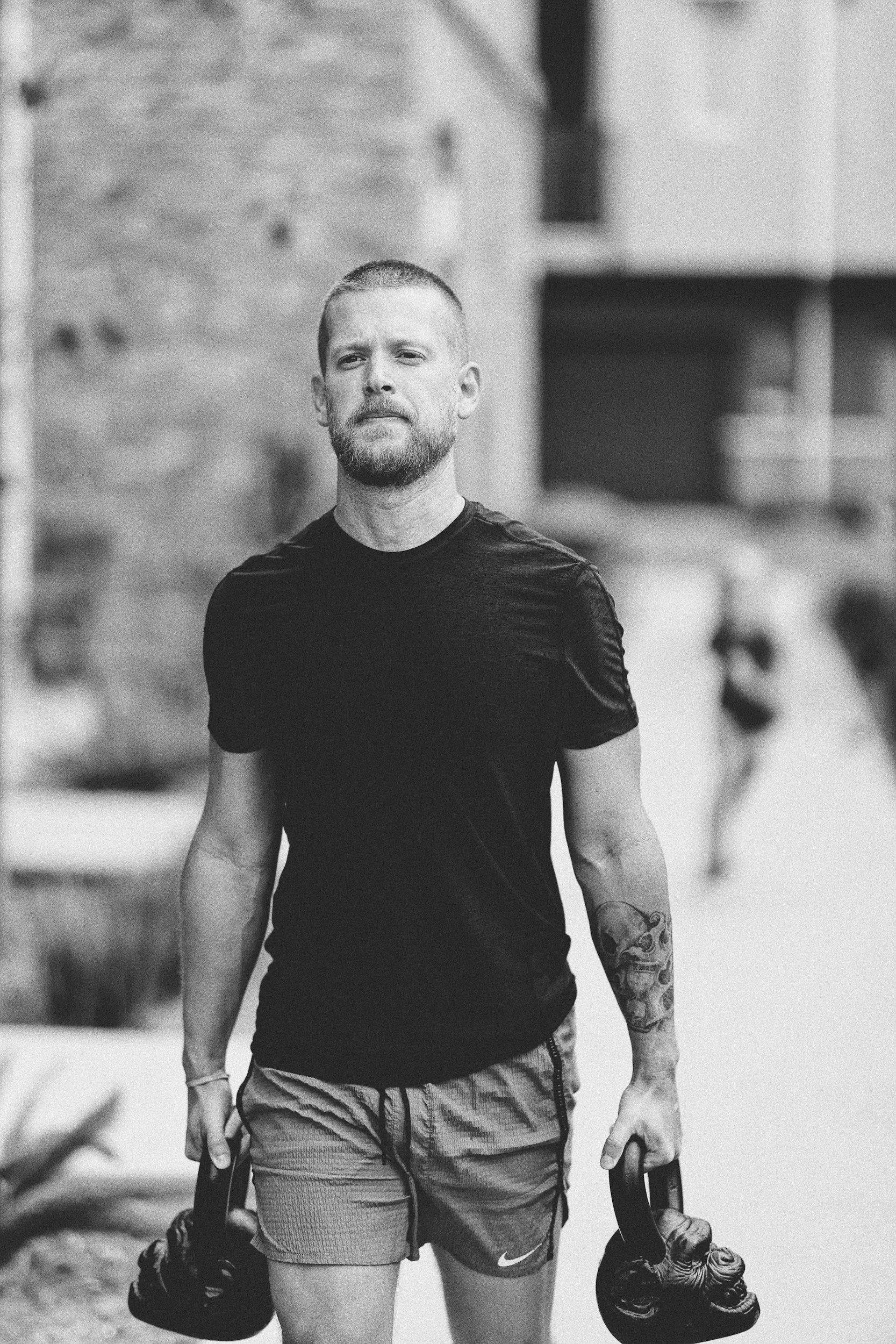 Man with short hair and beard walking outdoors, carrying kettlebells in both hands, wearing a black t-shirt and shorts, with a tattoo on his left forearm.