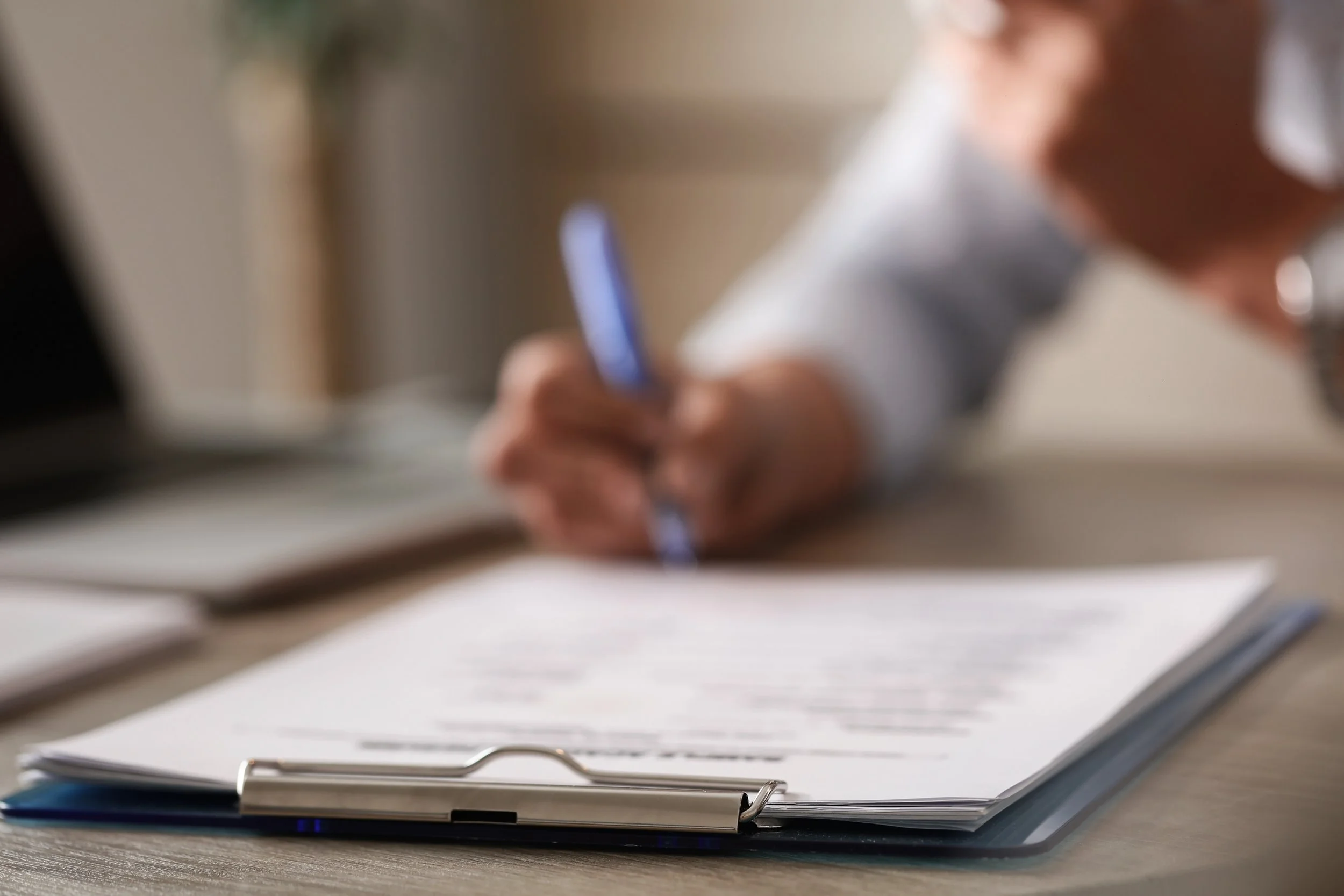 Blurred person writing on a document with a pen, with a clipboard holding papers on a desk