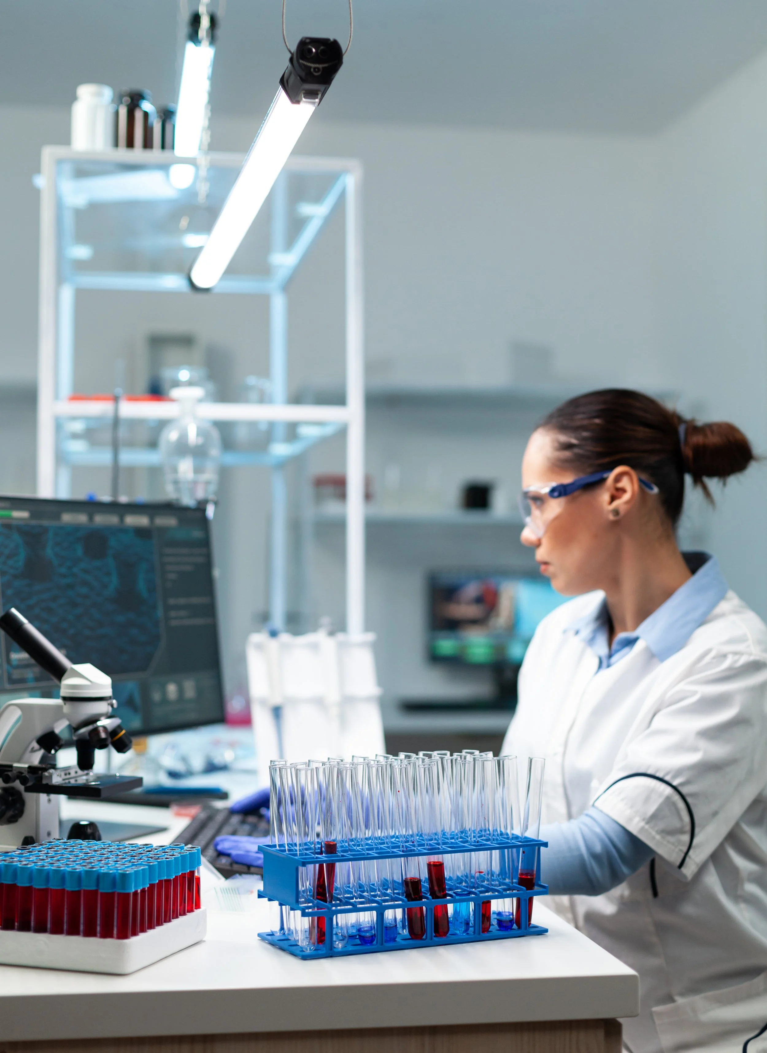 Laboratory scientist working with test tubes filled with blood samples, using a computer and microscope in a scientific lab.