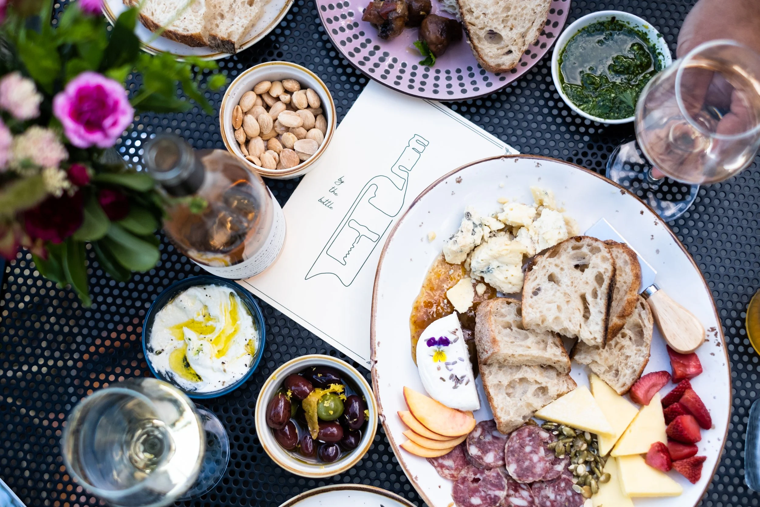 Salt and Bubbles' menu with food items on plates and bowls, including bread, cheese, fruit, nuts, and a charcuterie platter with meats and cheeses, on a black perforated table with glasses of white wine and a flower arrangement.