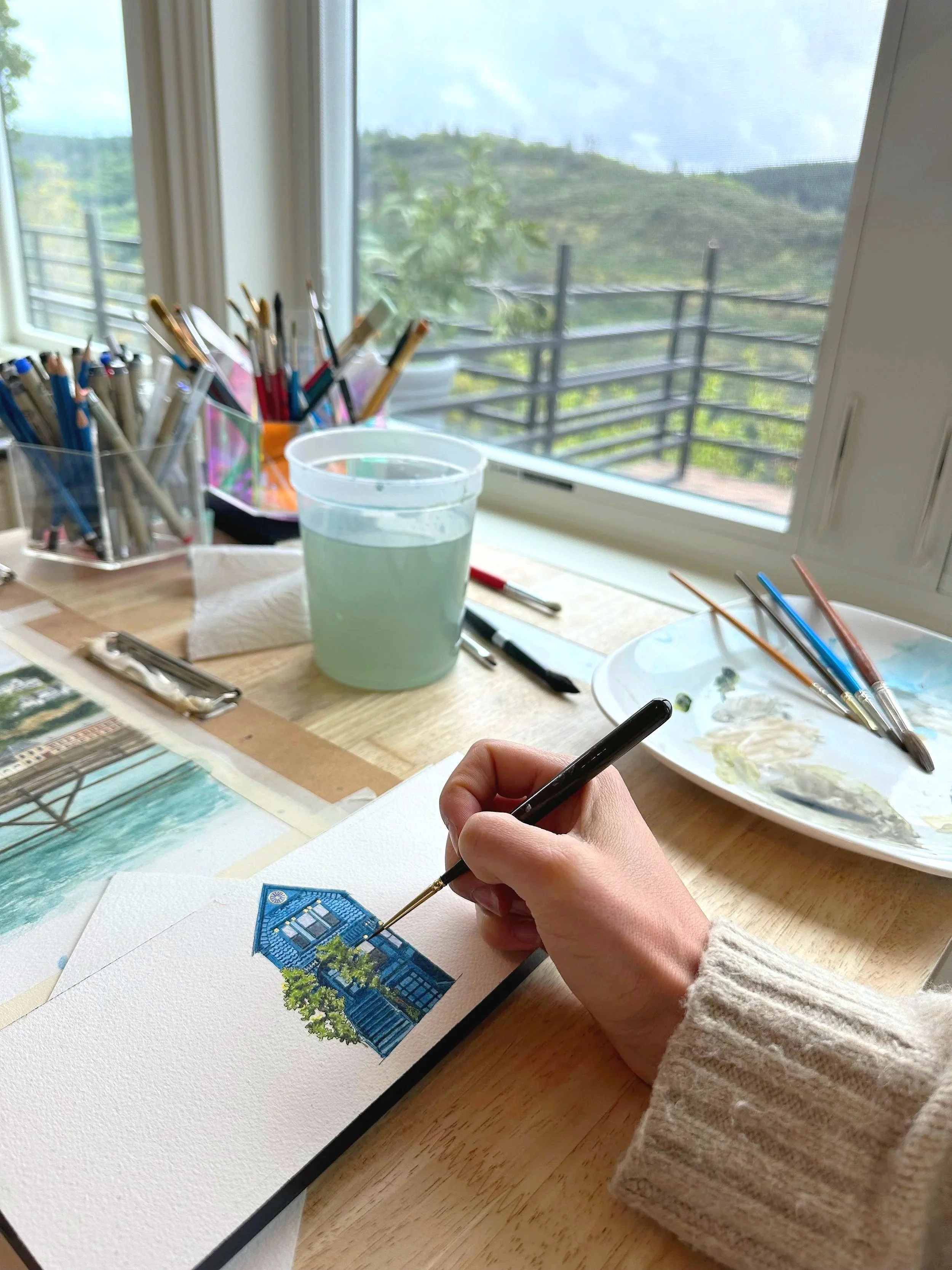 A person painting a detailed watercolor of a blue house with trees around it.